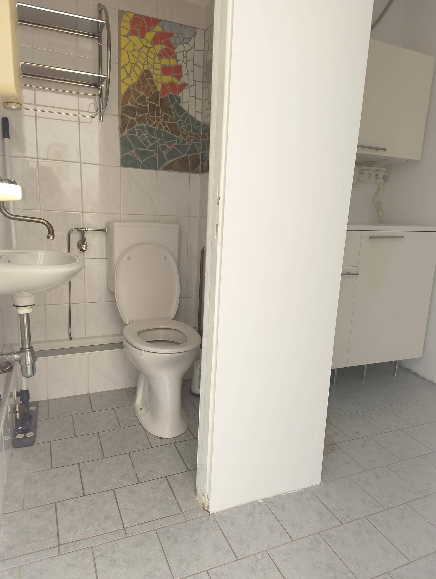Kleiner Toilettenraum mit WC, Waschbecken und weiß gefliesten Wänden sowie grauem Fliesenboden.