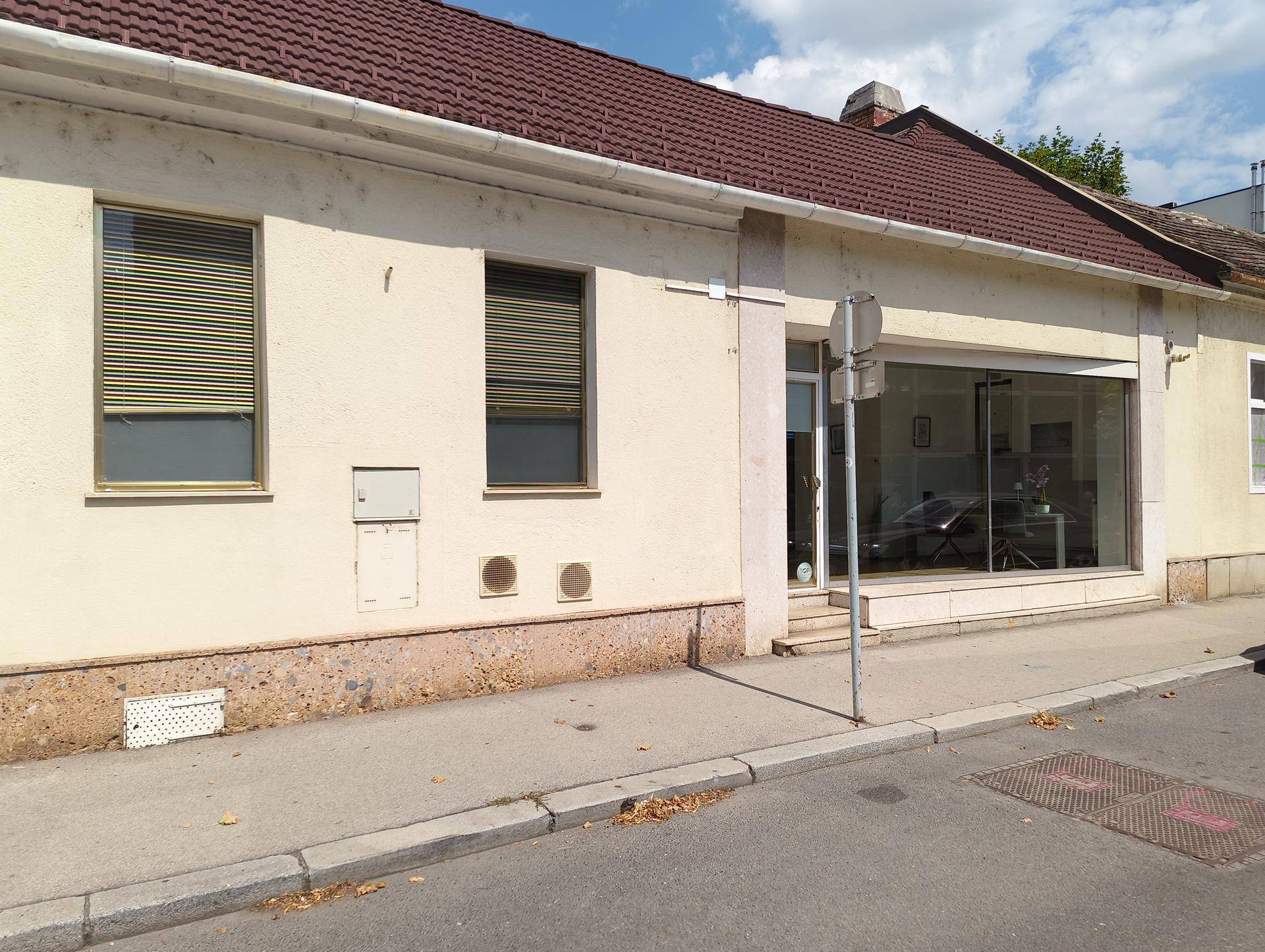 Gesamtansicht der Geschäftsfassade von der Straße aus mit Fenstern, Rollläden und dem gläsernen Eingangsbereich.