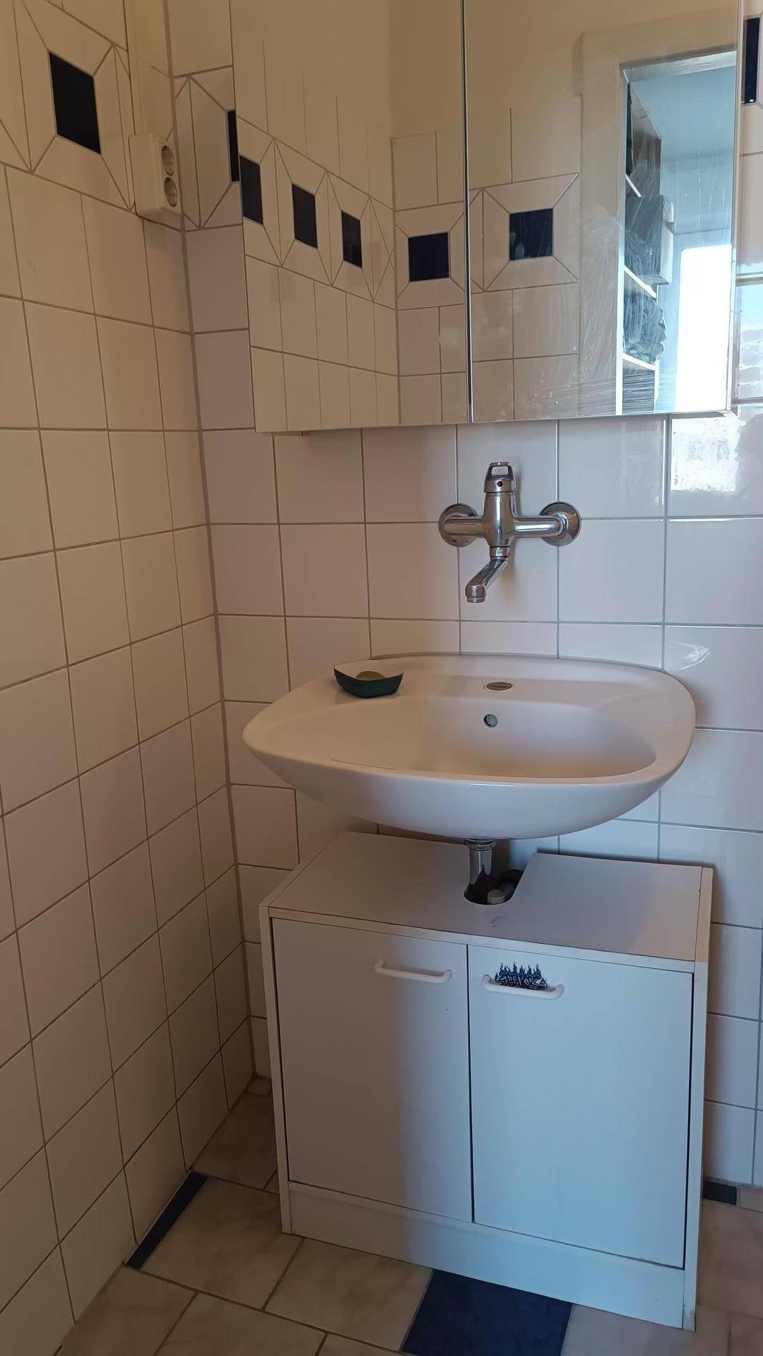 Badezimmer mit weiß gefliesten Wänden, Einzelwaschbecken und Spiegelschrank.
