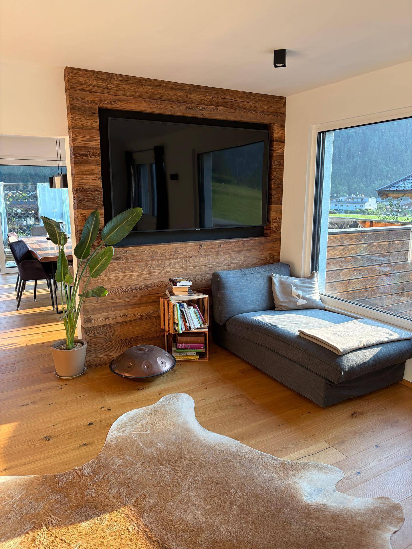 Gemütlicher Wohnbereich mit Chaiselongue, großem Fernseher und Blick auf die umliegende Berglandschaft.