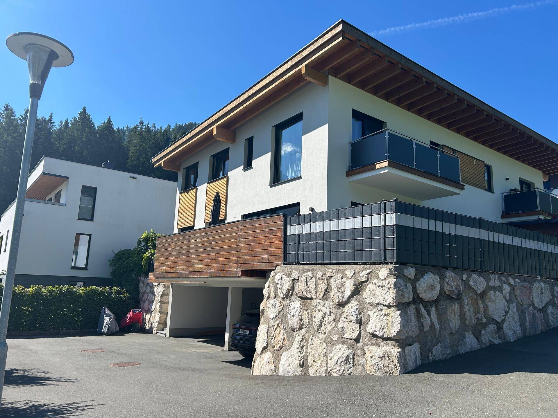 Modernes Haus mit weißer Fassade, Holzelementen, Balkonen und integriertem Carport vor Bergkulisse.