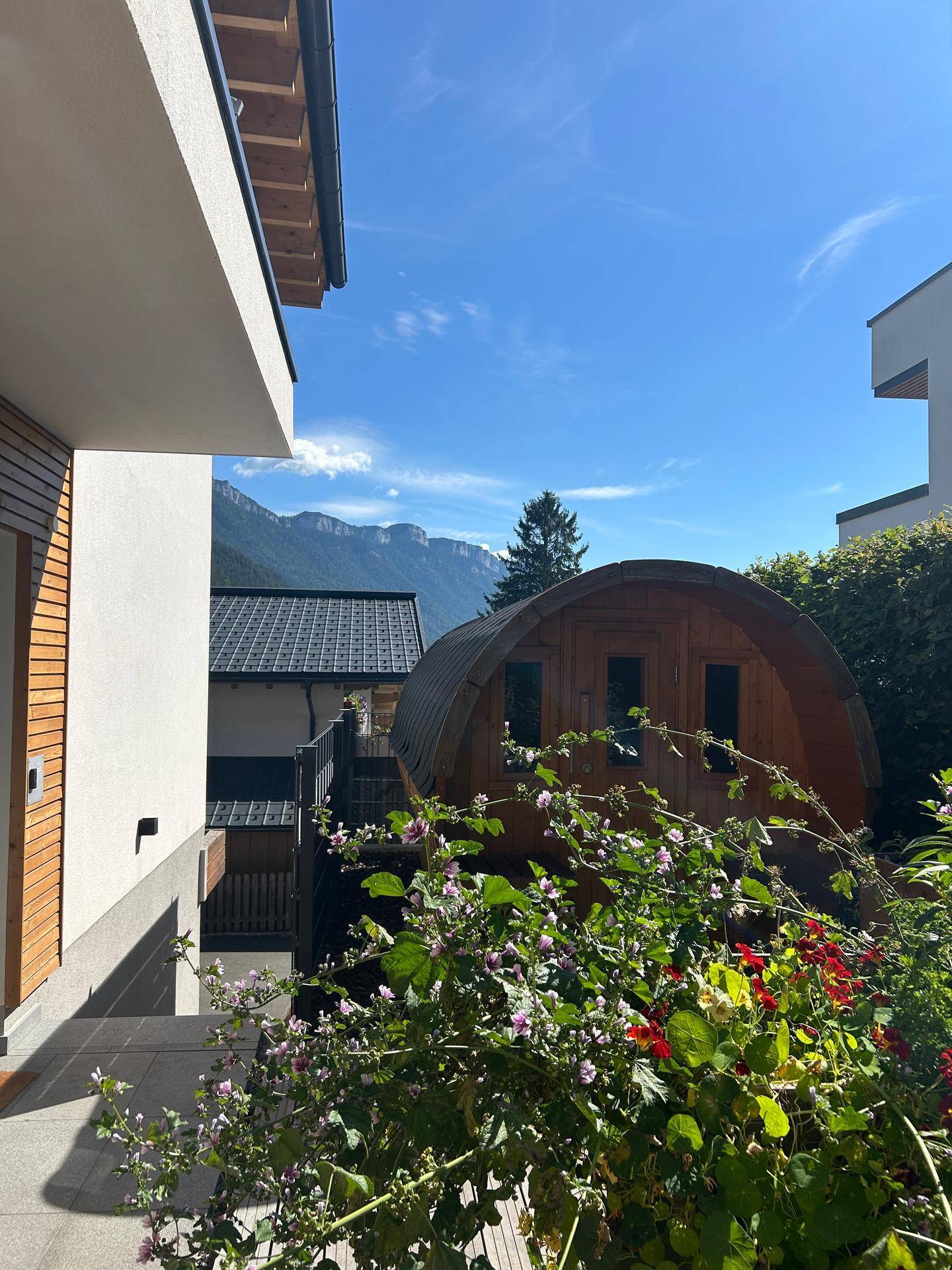 Außenansicht der Fasssauna im Gartenbereich mit blühenden Pflanzen und majestätischem Bergblick.