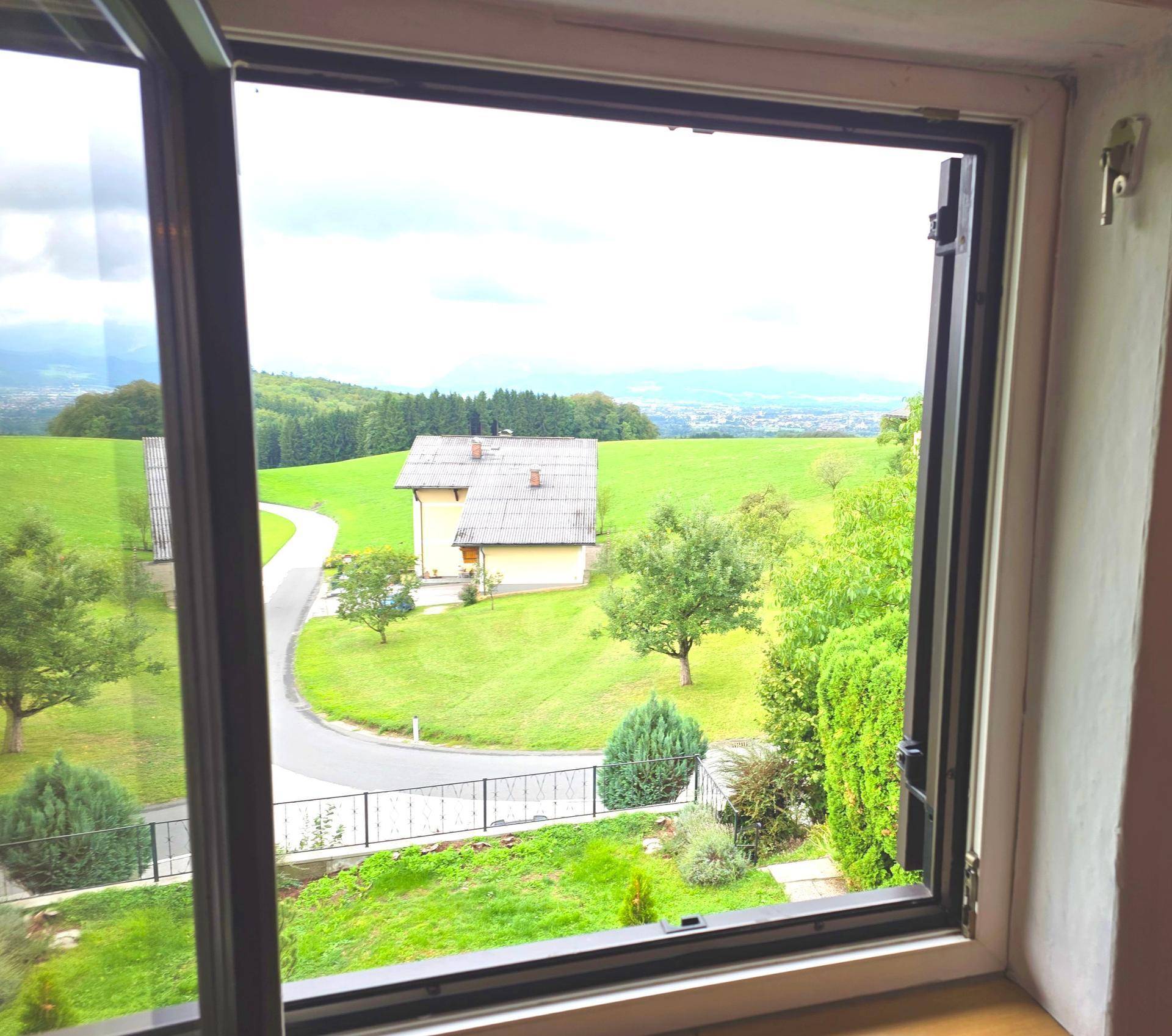 Weitblick aus dem Fenster über eine grüne Hügellandschaft mit Häusern und Bergen am Horizont.