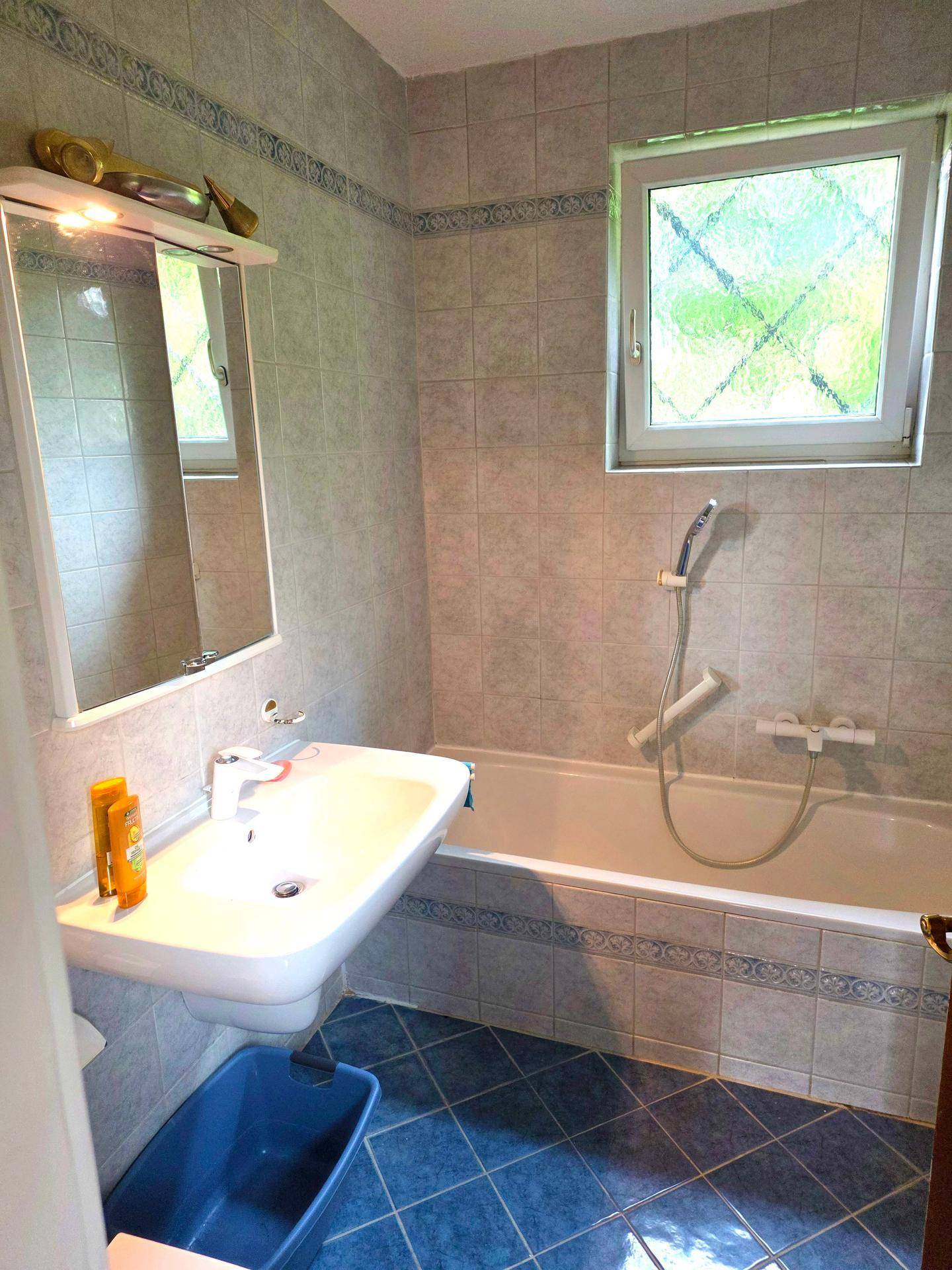 Badezimmer mit Badewanne, Waschbecken und hellblauen Fliesen, beleuchtet durch ein Fenster.