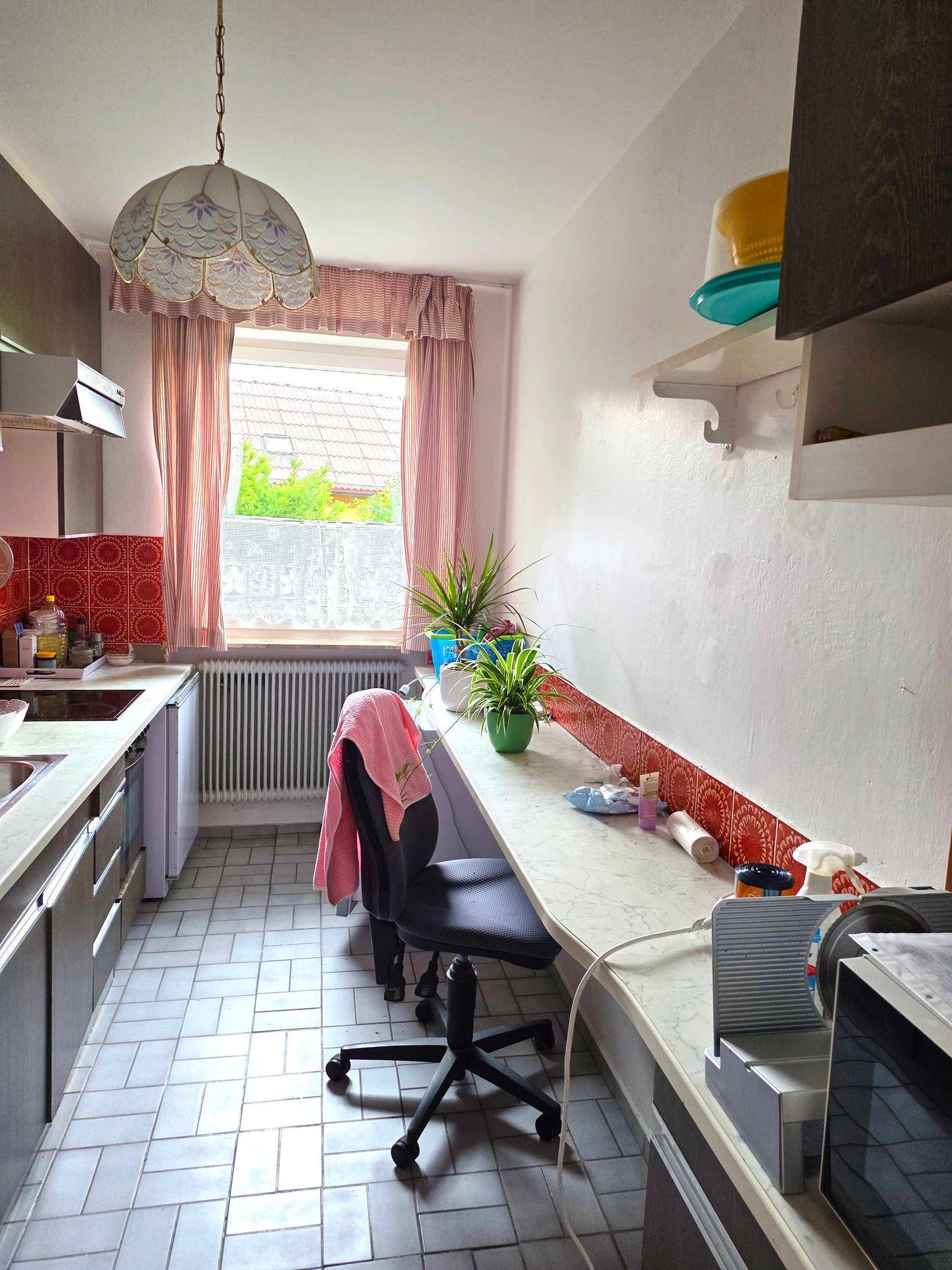 Schmale Küche mit Fliesenboden, Arbeitsplatte und Fenster mit rosa Vorhängen und Heizkörper.
