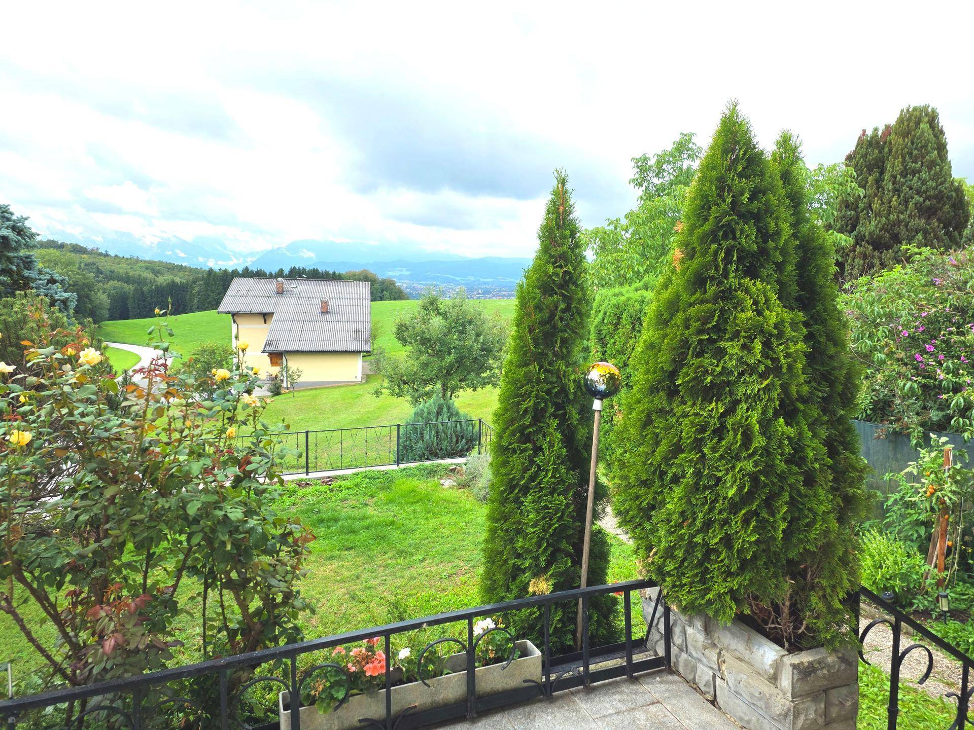 Terrasse mit Blick auf die grüne Umgebung, einen Garten und entfernte Berge unter bewölktem Himmel.