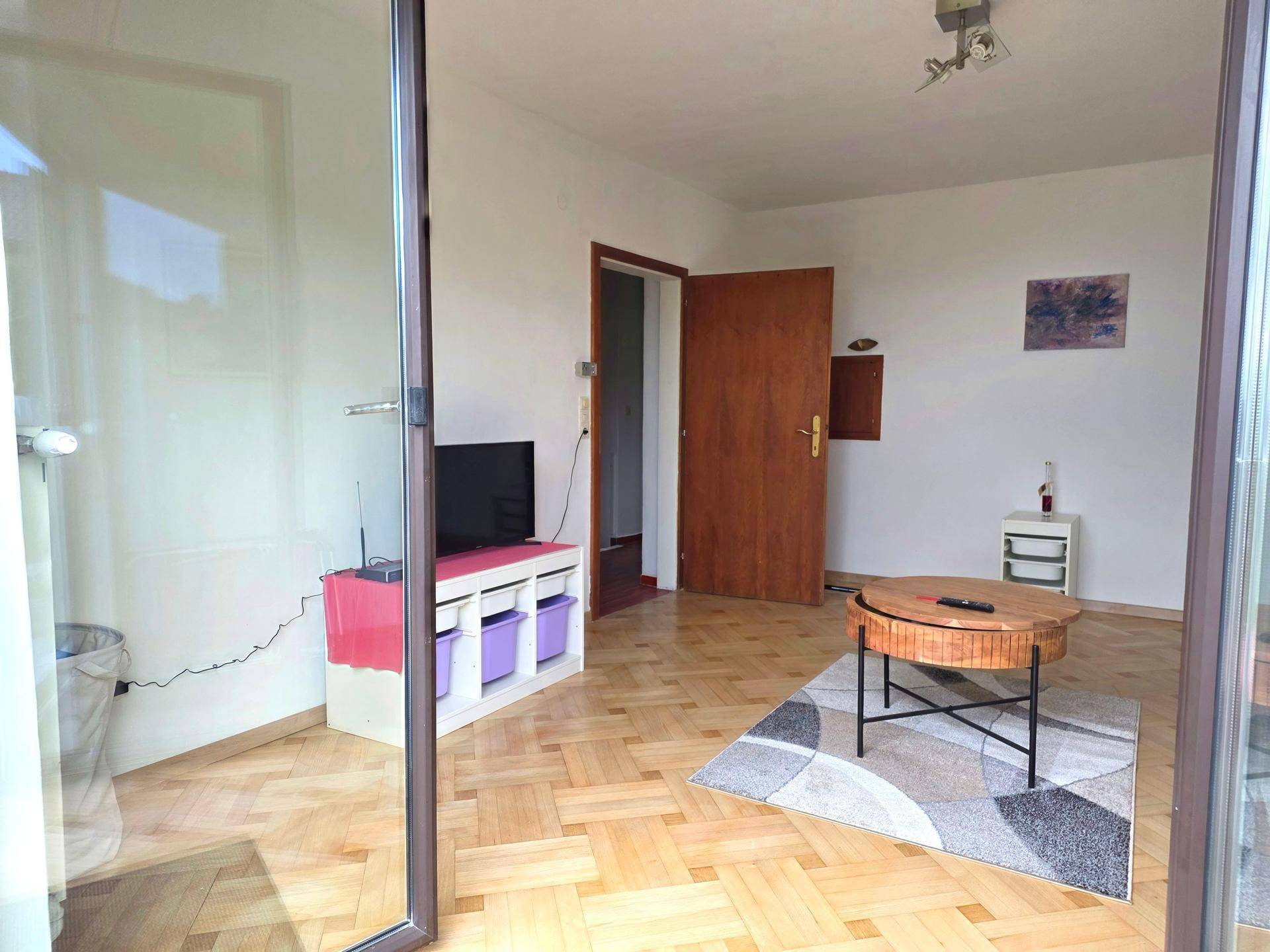 Wohnzimmer mit Parkettboden, Fernseher und einem runden Couchtisch, durch eine offene Tür.