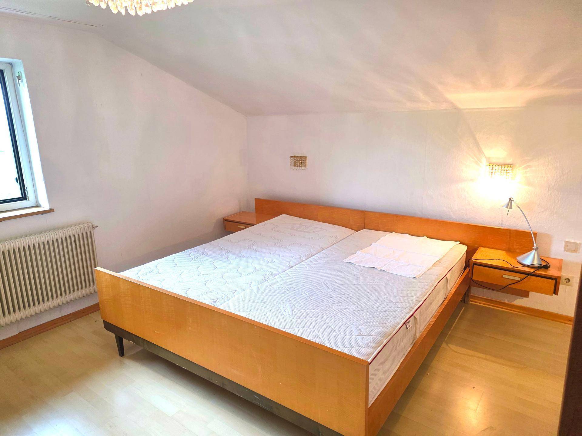 Schlafzimmer mit Doppelbett, Holzboden und Dachschräge, hell beleuchtet durch ein kleines Fenster.