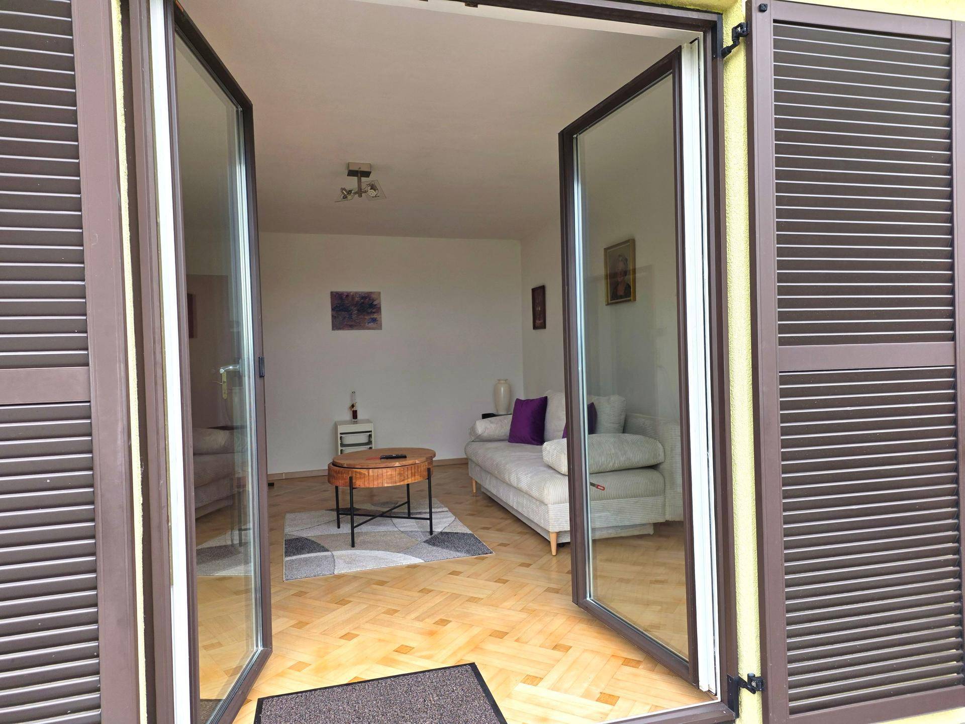 Wohnzimmer mit Parkettboden und Sofa, gesehen durch weit geöffnete Glastüren mit Fensterläden.