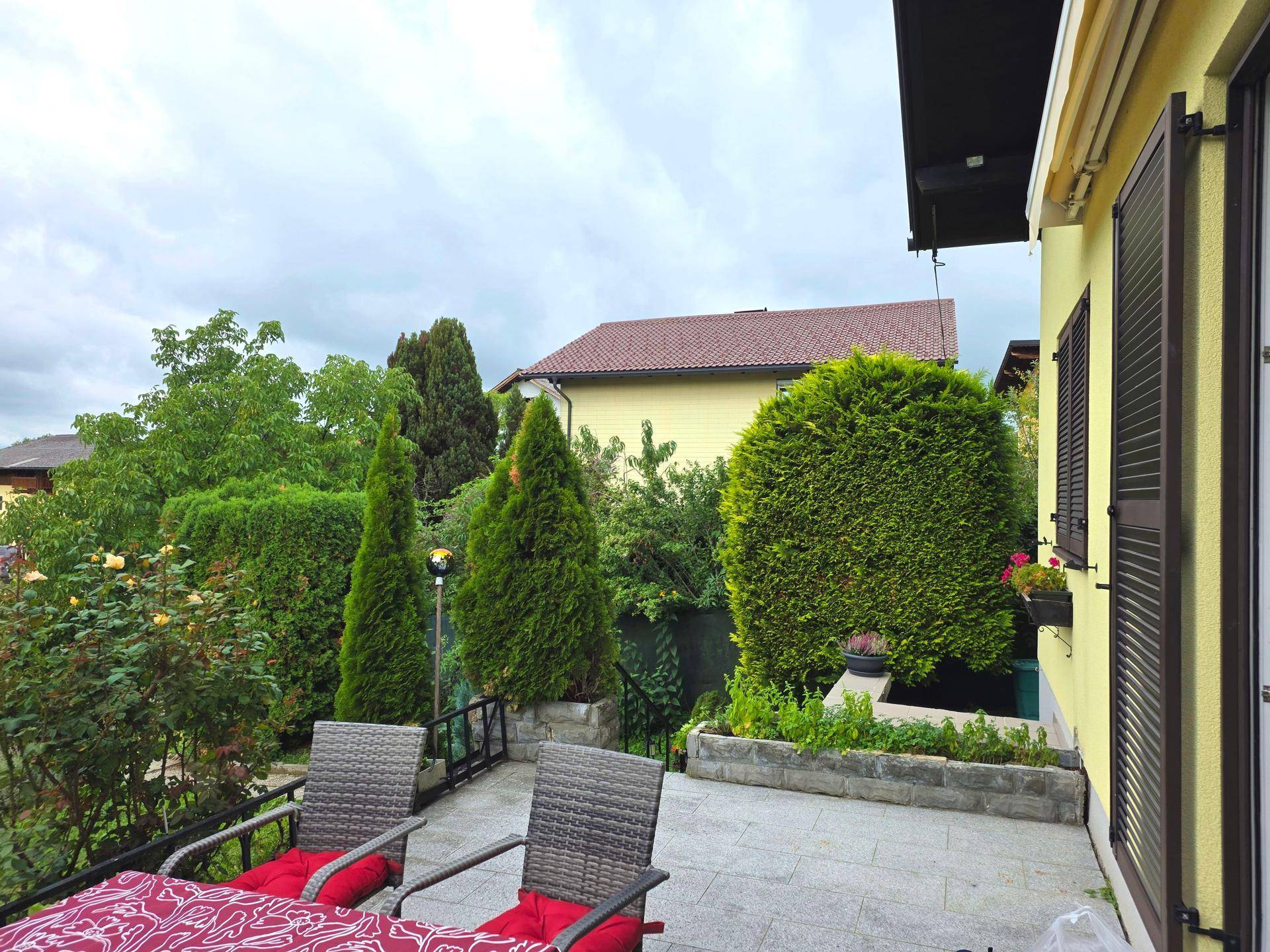 Terrasse mit Sitzgelegenheiten neben dem Haus, umgeben von hohen Hecken und Bäumen im Garten.