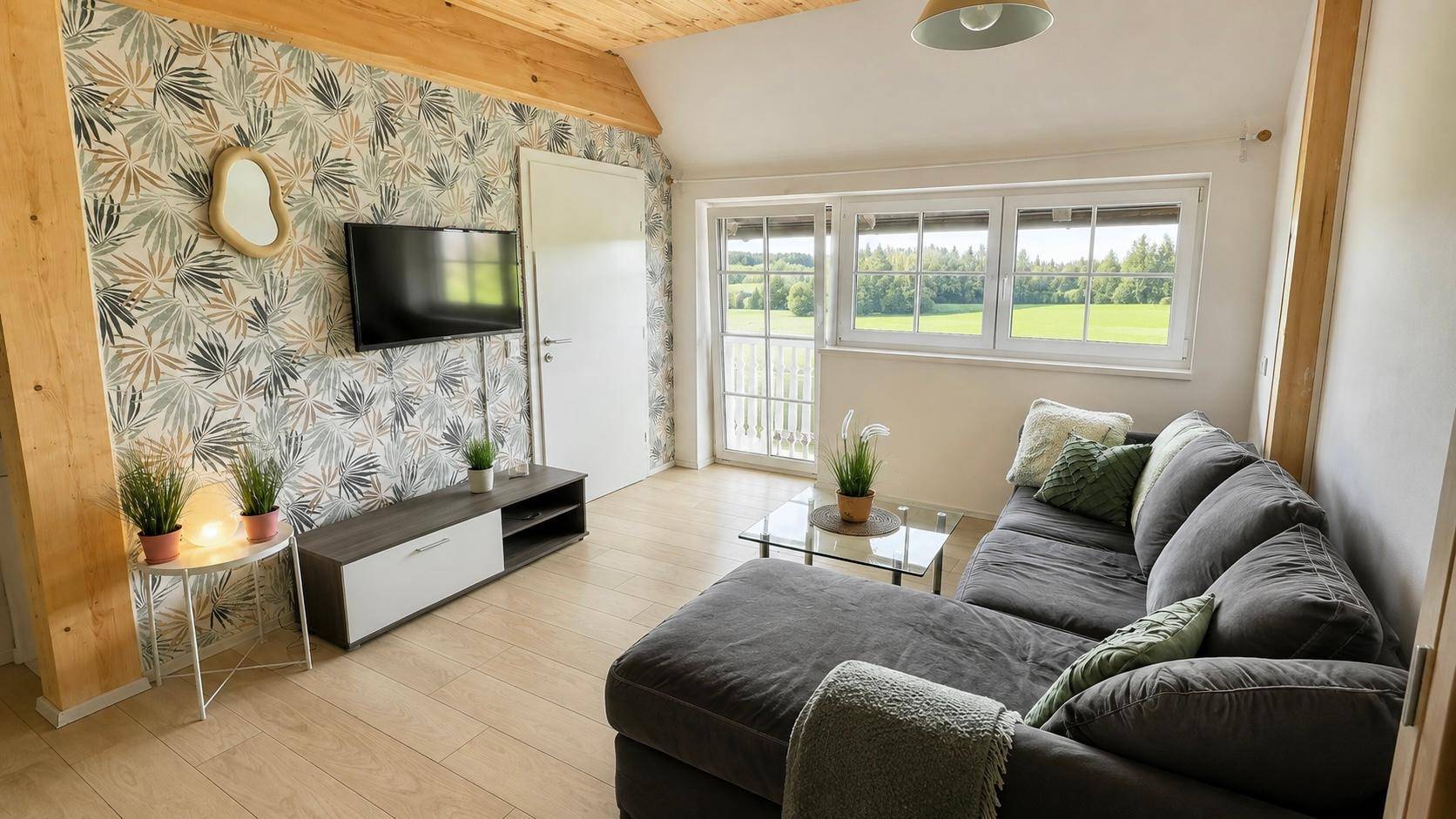 Helles Wohnzimmer mit gemütlichem Sofa, Fernseher und Blick auf die grüne Landschaft.