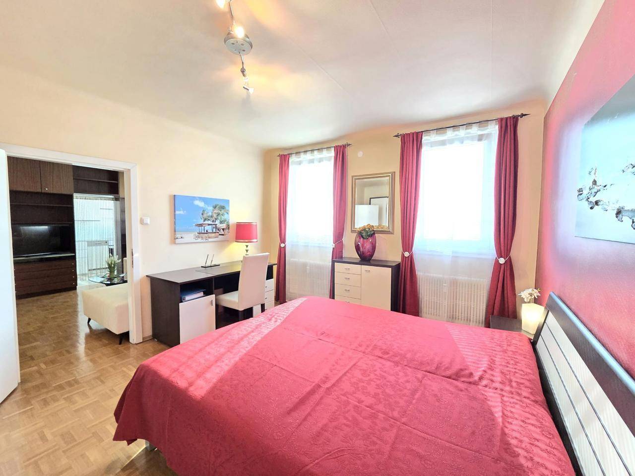 Schlafzimmer mit Doppelbett, Schreibtisch und Parkettboden, hell durch große Fenster.