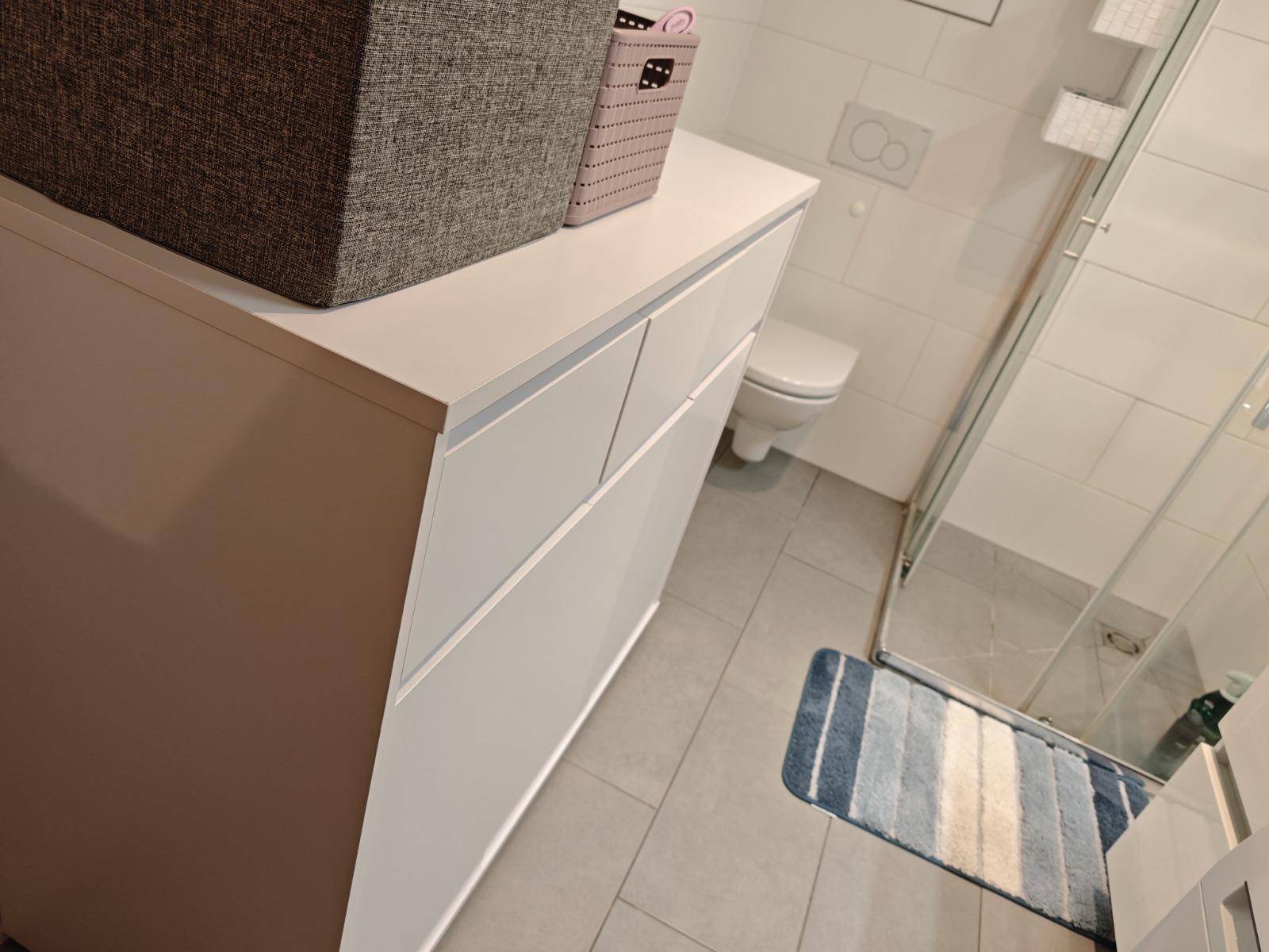 Modernes Badezimmer mit hellgrauen Fliesen und weißem Schrank, Toilette im Hintergrund.
