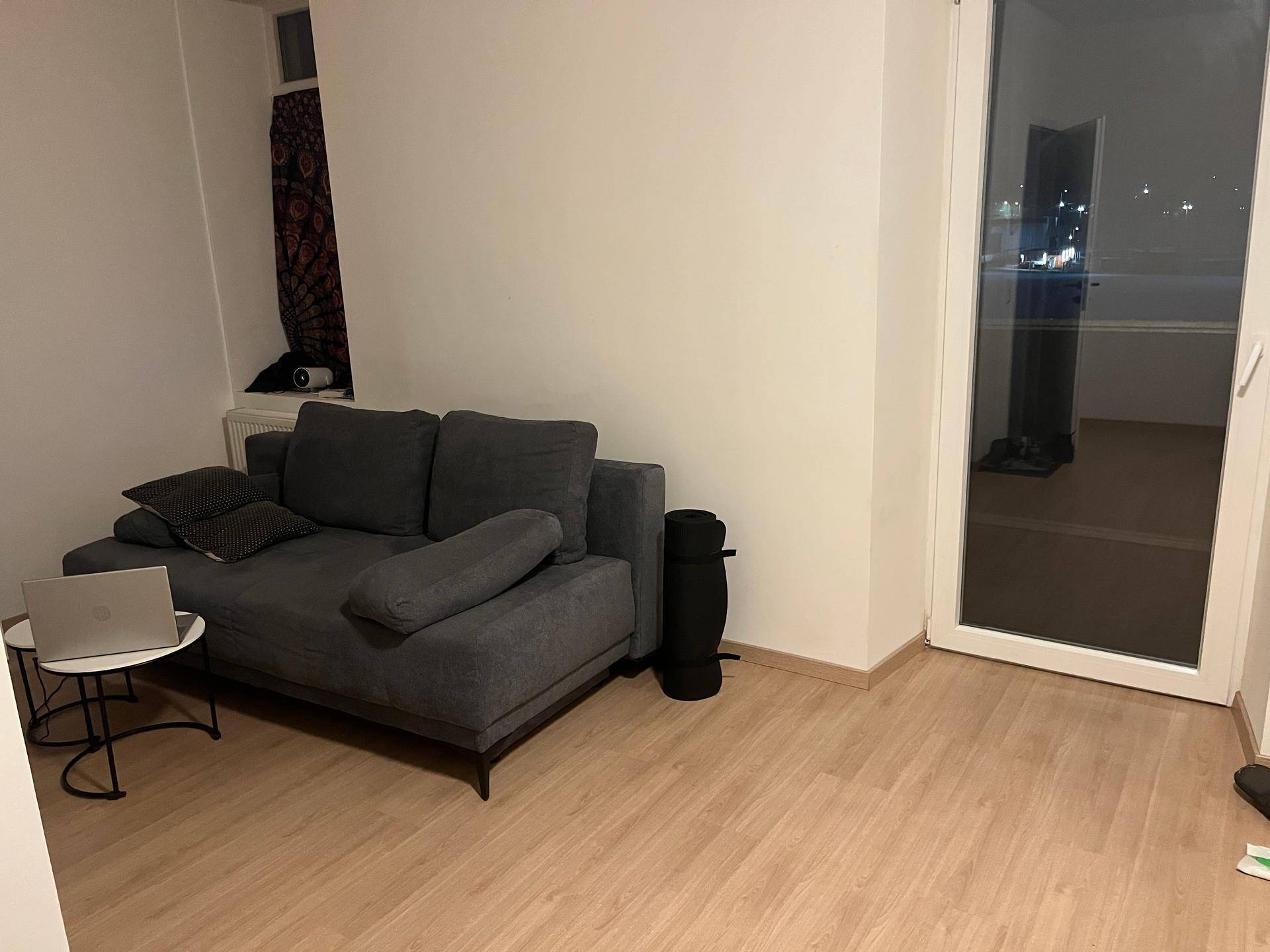 Wohnzimmer mit einem grauen Sofa, Laptop und Zugang zu einem Balkon oder einer Terrasse.