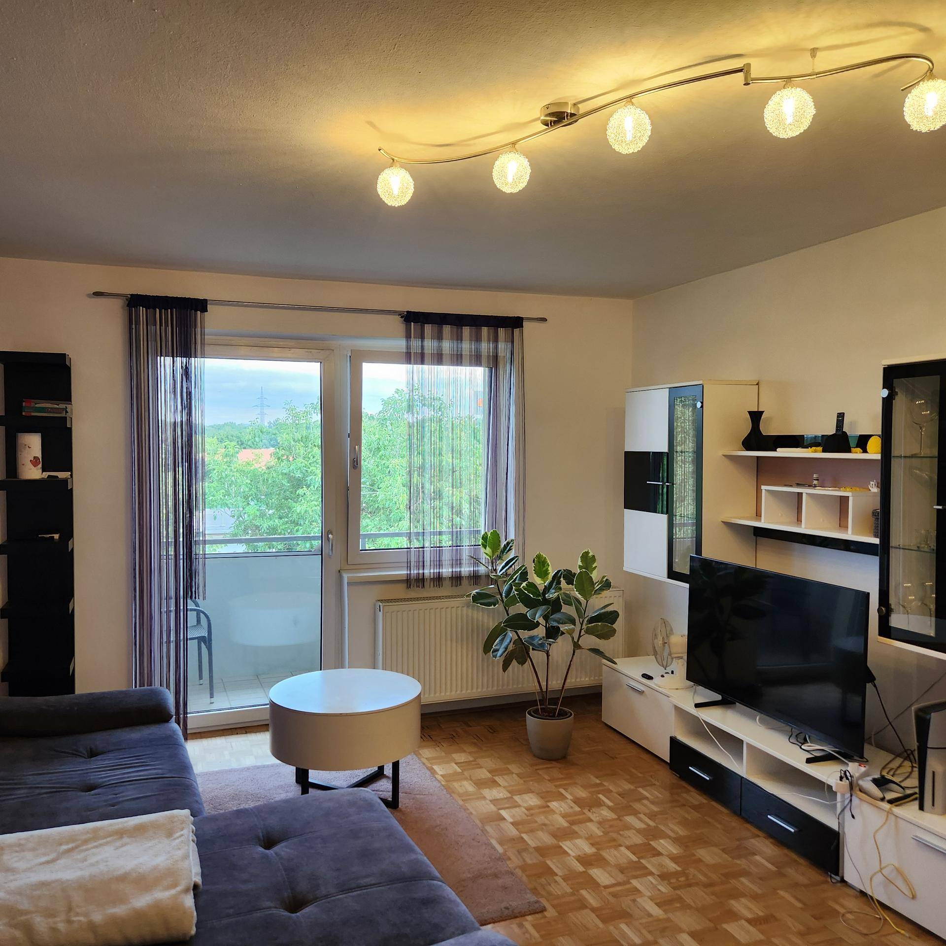 Gemütliches Wohnzimmer mit Sofa, Fernseher und Zugang zum Balkon mit Blick ins Grüne.
