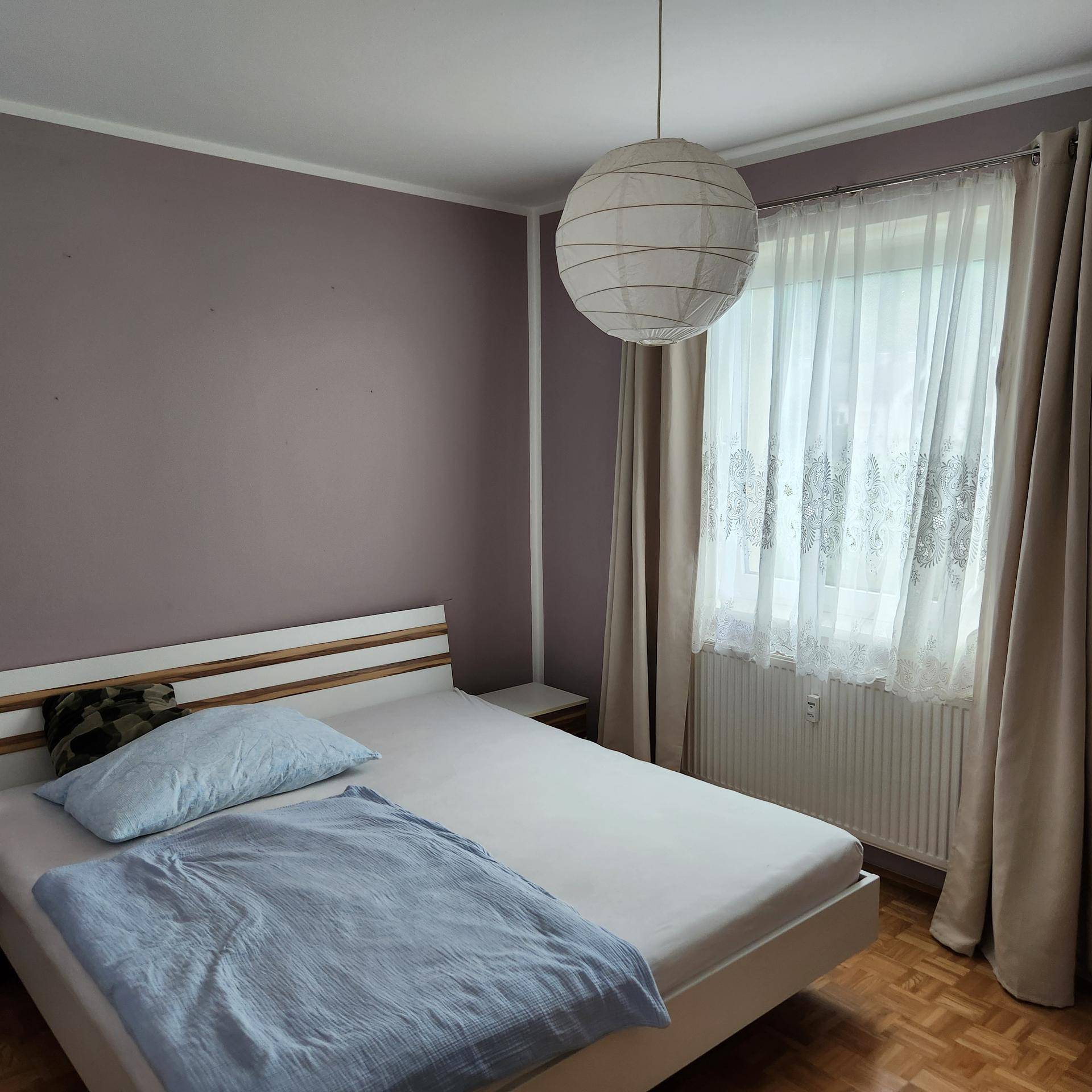 Schlafzimmer mit Doppelbett, Nachttischen und einem Fenster mit hellen Vorhängen für Tageslicht.
