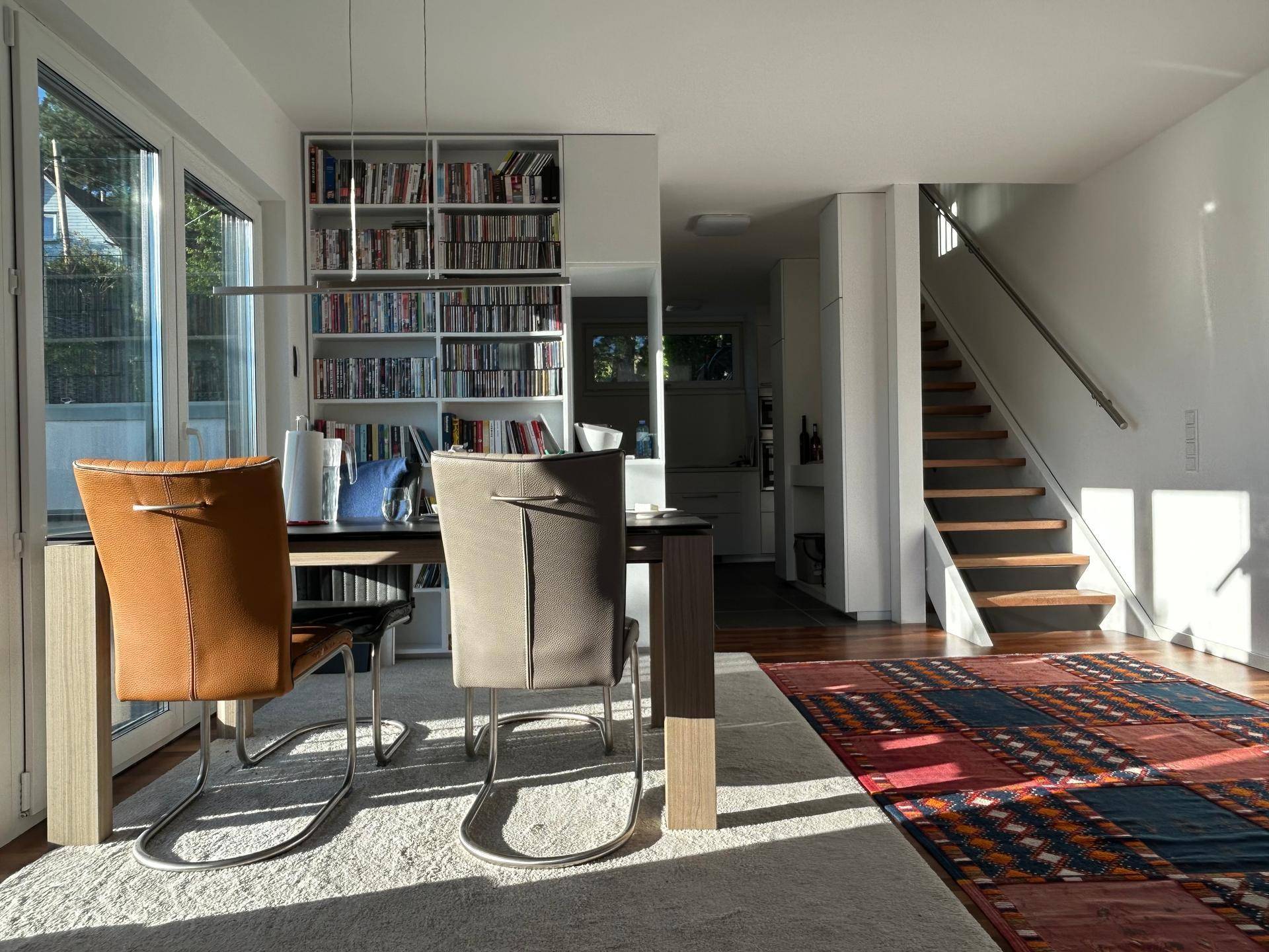 Heller Wohn-Essbereich mit Holzboden, integriertem Bücherregal und offener Treppe.