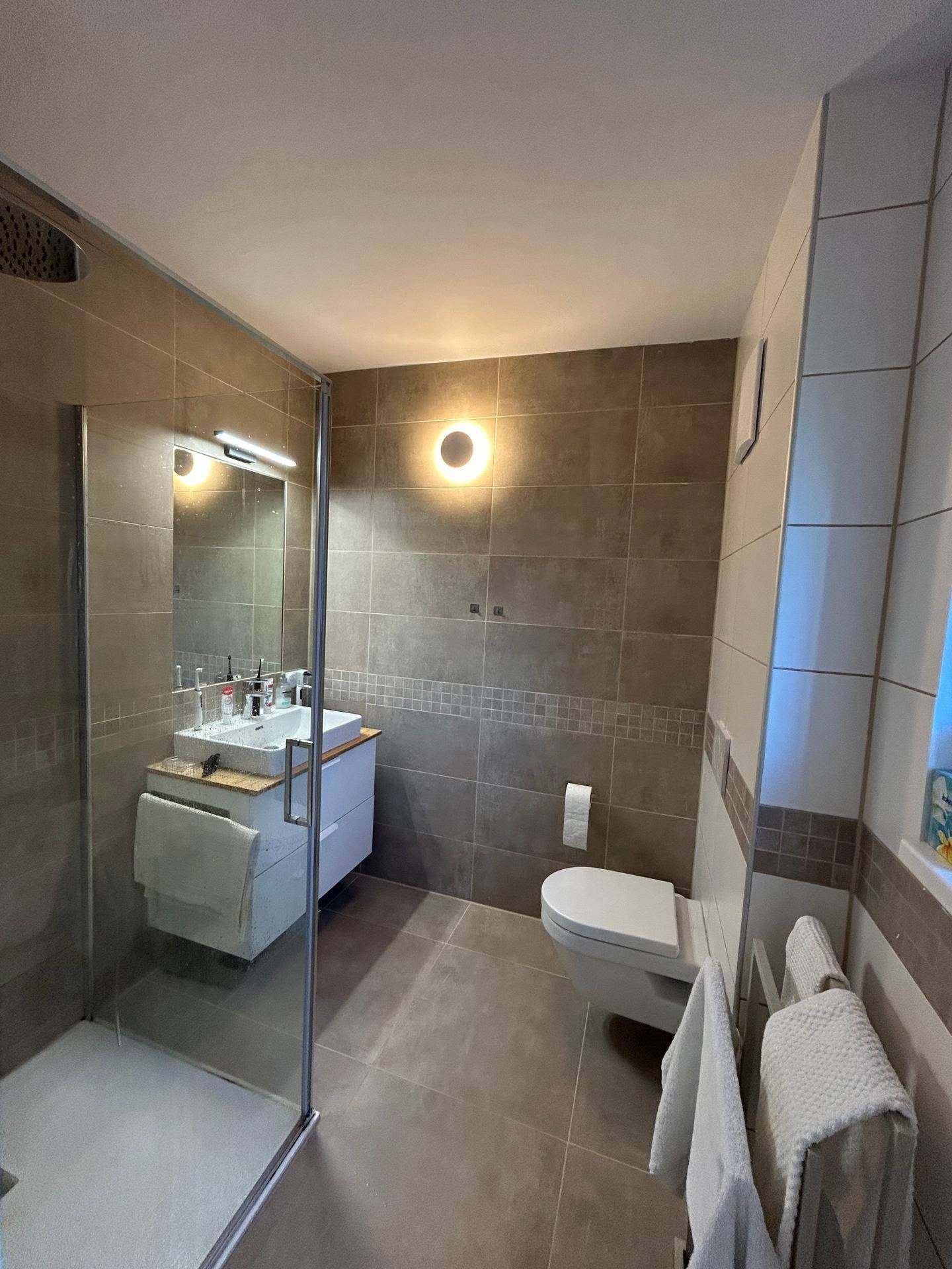 Modernes Badezimmer mit grauen Fliesen, begehbarer Dusche und wandhängendem WC.