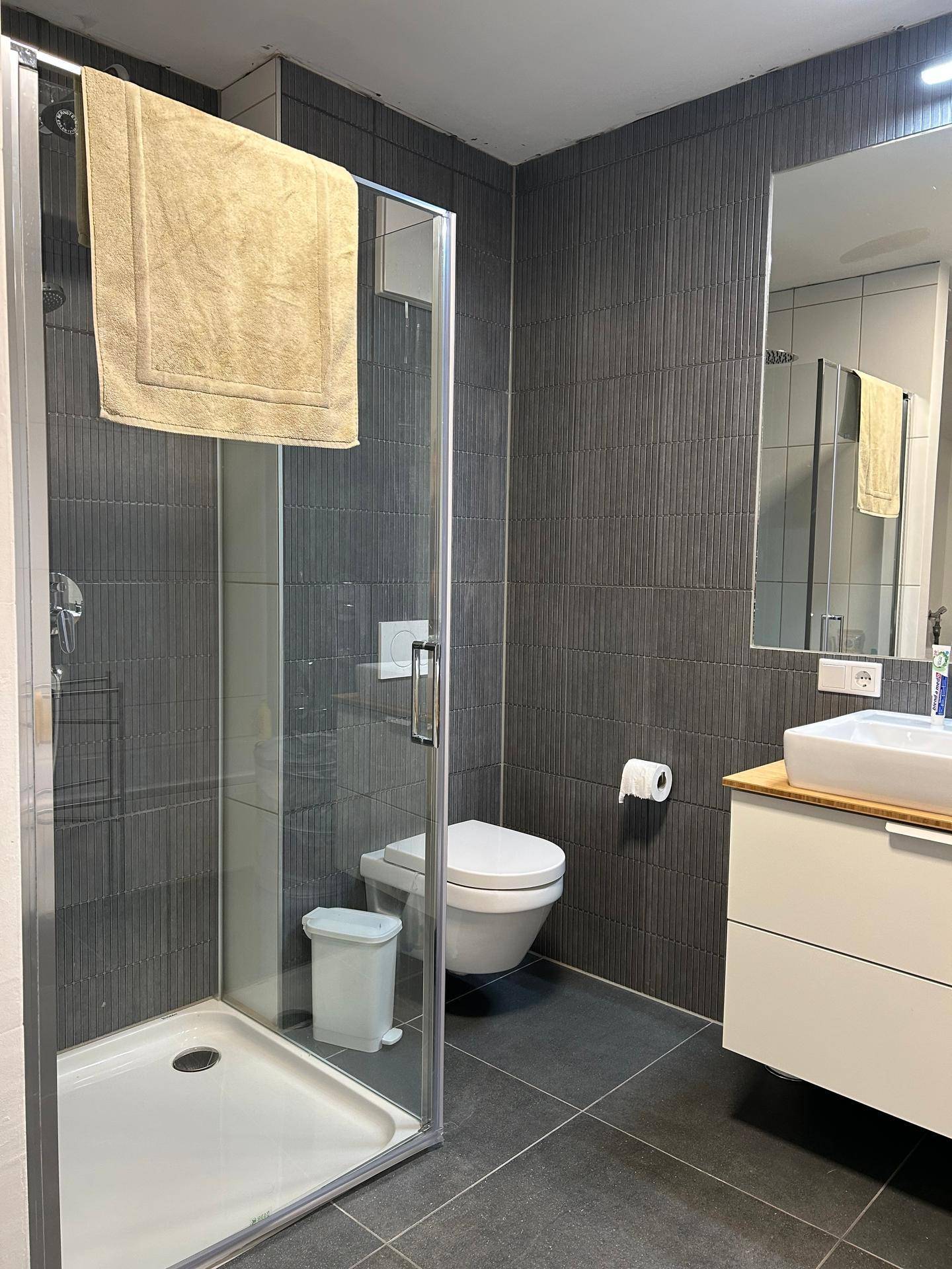 Modernes Badezimmer mit dunklen Fliesen, begehbarer Dusche und minimalistischem Waschtisch.