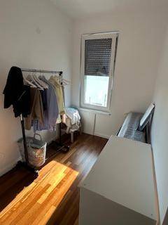 Kleines Zimmer mit Holzboden, Kleiderständer und Fenster mit Rollladen.
