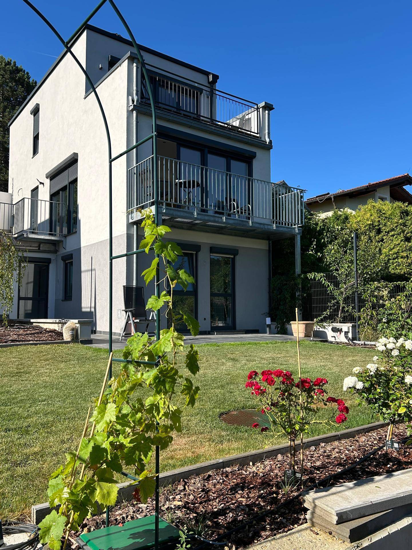 Moderne Hausfassade mit mehreren Balkonen und gepflegtem Garten unter blauem Himmel.