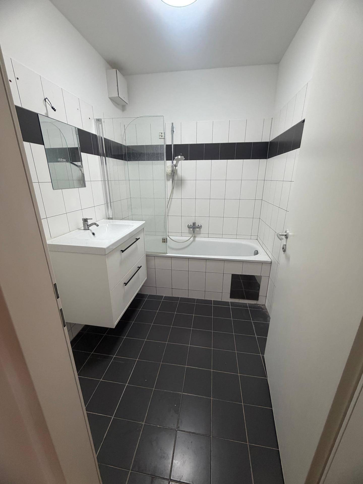 Badezimmer mit weißen Wandfliesen, dunklem Boden, Badewanne mit Duschfunktion und Waschtisch.