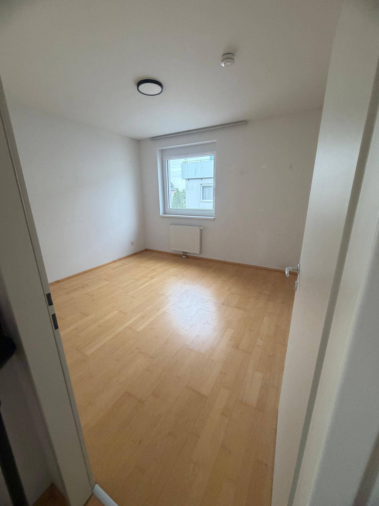 Leeres Schlafzimmer mit hellem Parkettboden und einem Fenster, das Tageslicht hereinlässt.