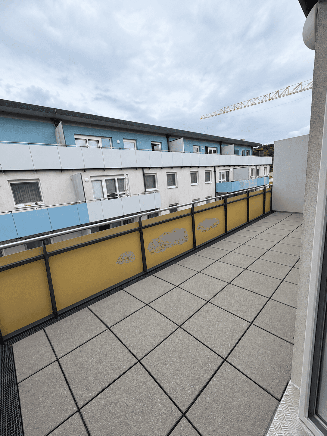 Geräumiger Balkon mit Fliesenboden und gelbem Geländer, Blick auf gegenüberliegende Gebäude.