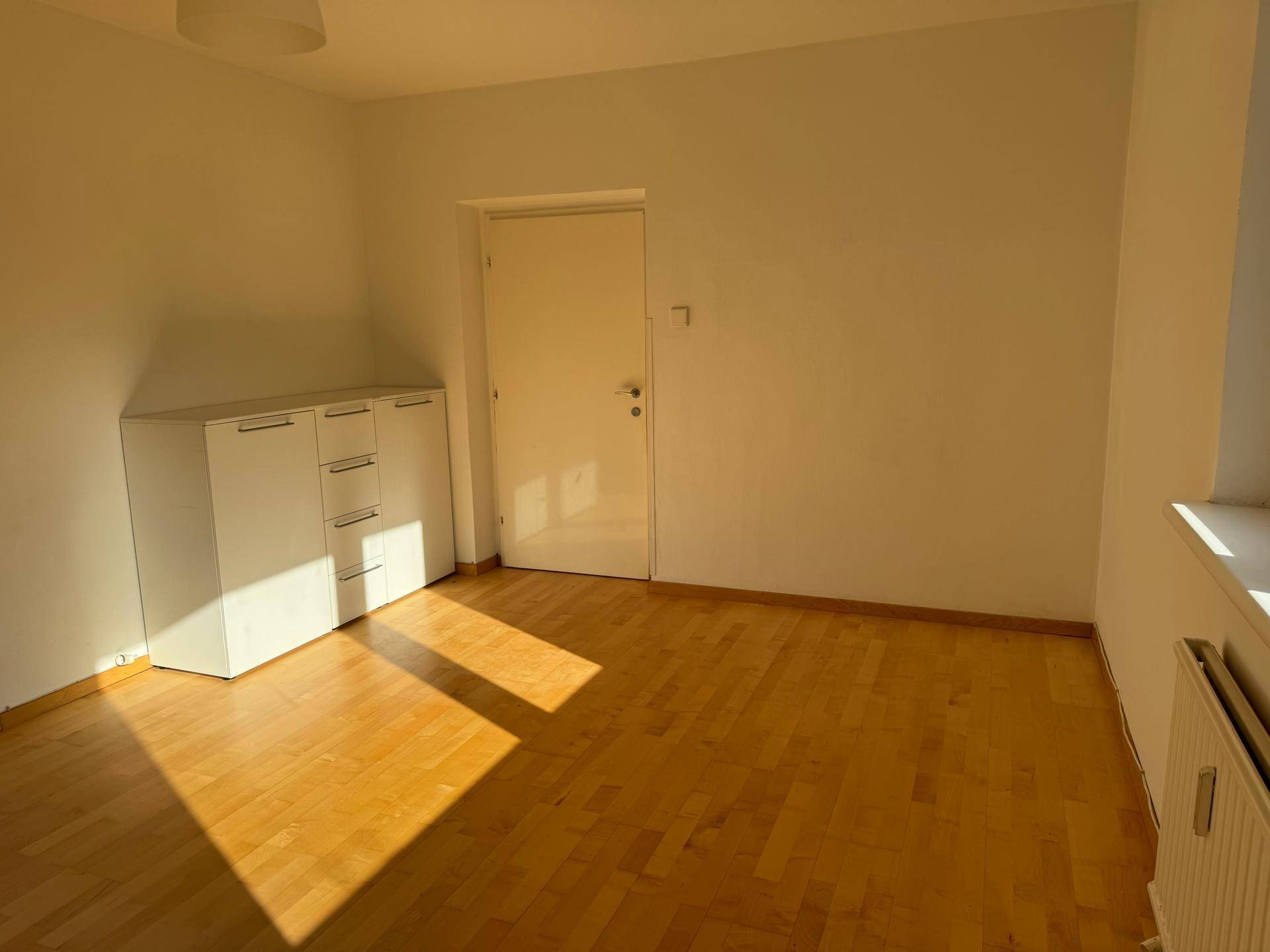 Heller Raum mit Parkettboden, weißer Kommode und viel Sonnenlicht.