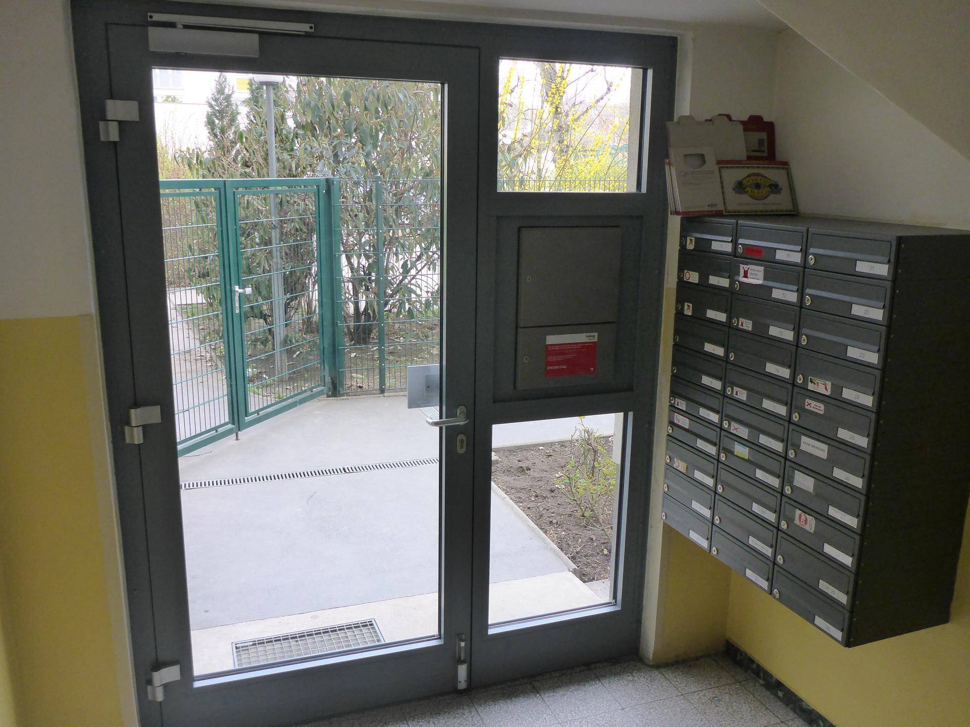 Eingangsbereich eines Mehrfamilienhauses mit Briefkästen und Blick auf einen umzäunten Außenbereich.