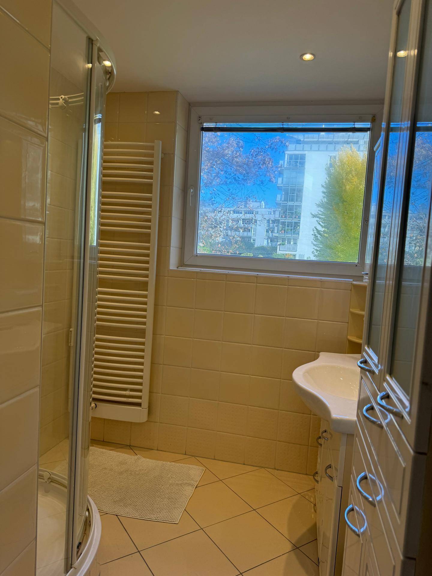 Badezimmer mit beigen Fliesen, Duschkabine, Waschbecken und Fenster.