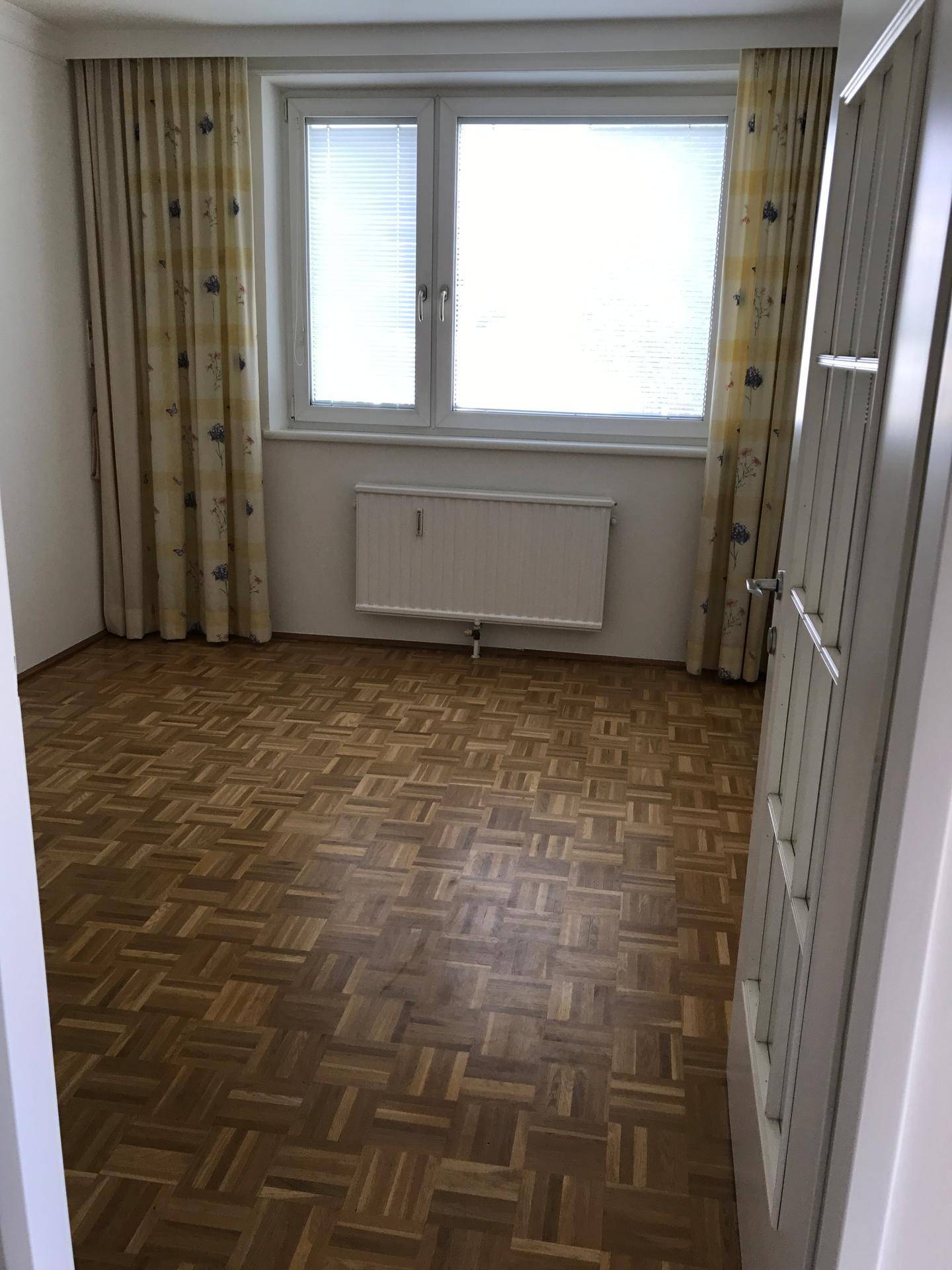 Helles Schlafzimmer mit Fischgrätparkett, Fenster mit Jalousien und gemusterten Vorhängen.