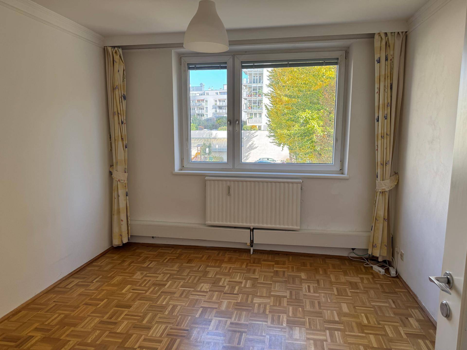 Leerer Raum mit Parkettboden, Fenster mit Vorhängen und Blick auf die Umgebung.