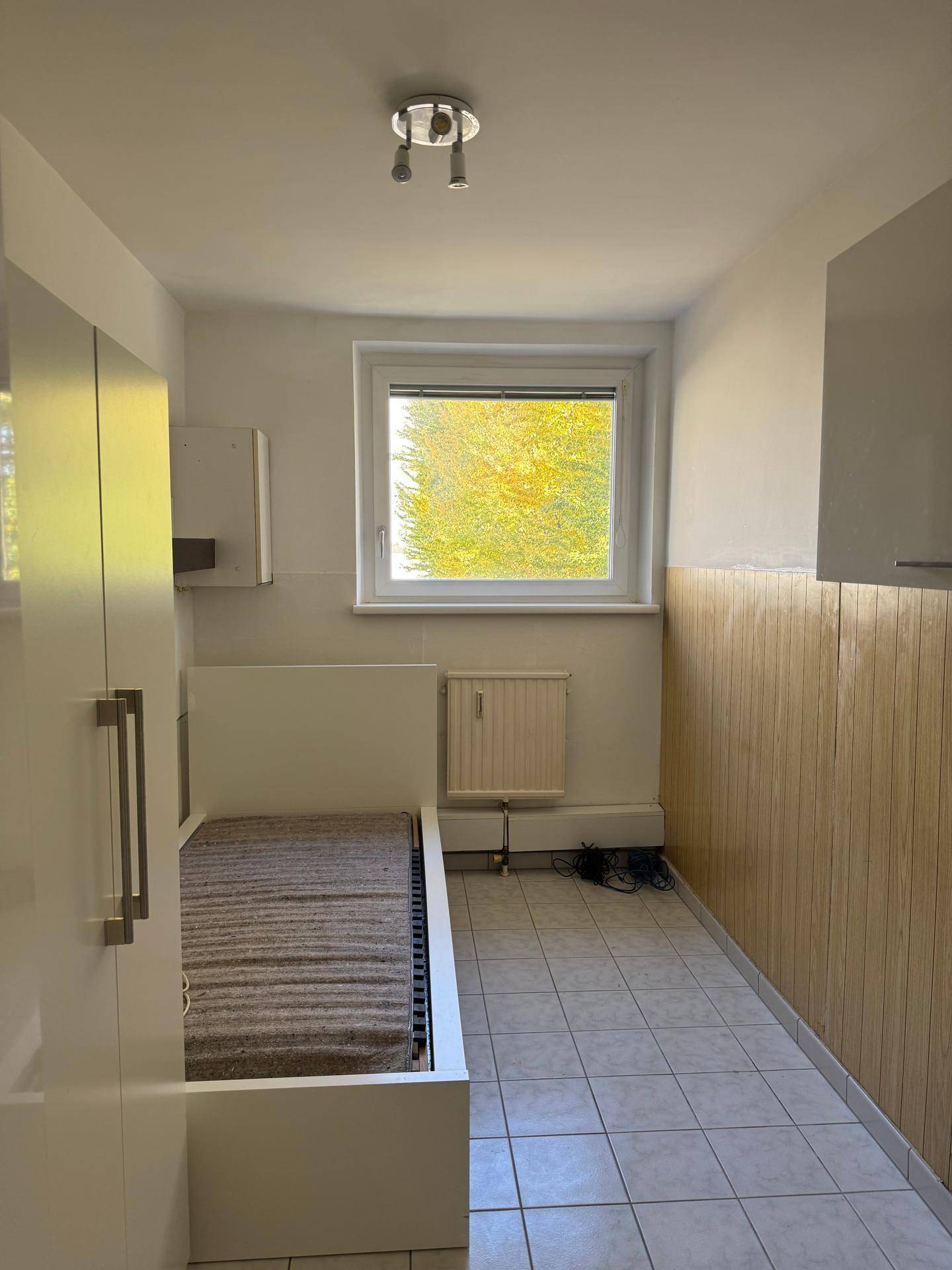 Kleines Schlafzimmer mit Fliesenboden, Einzelbett und Blick auf grüne Bäume.