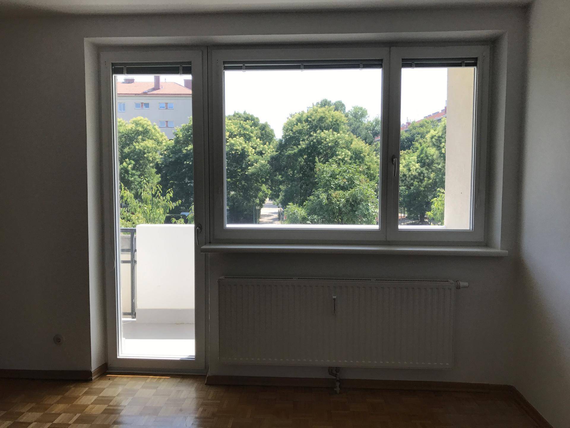 Heller Raum mit Parkettboden, großem Fenster und Zugang zu einem Balkon mit Blick ins Grüne.