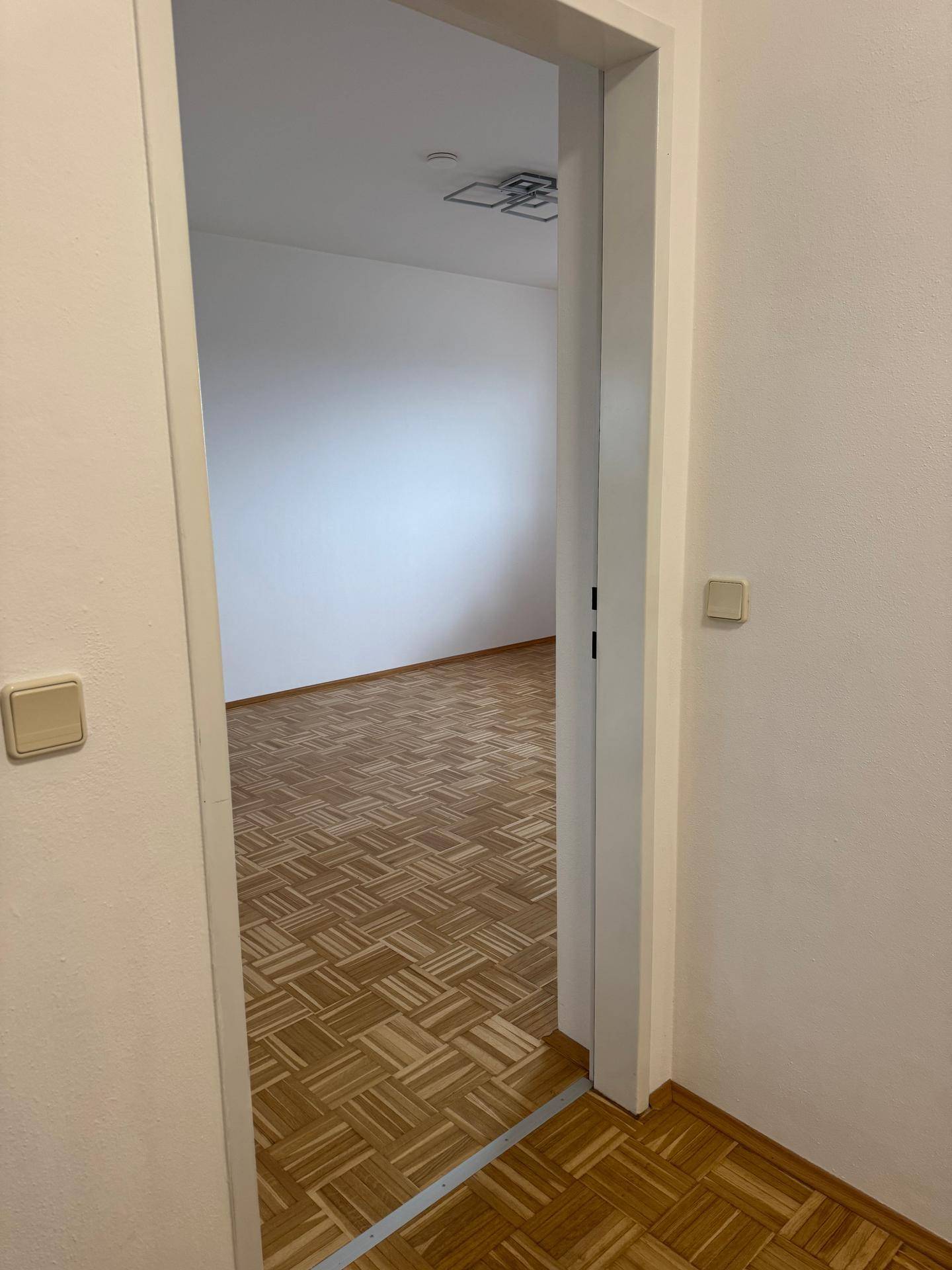 Blick durch eine Tür in einen leeren Raum mit hellem Parkettboden und weißen Wänden.