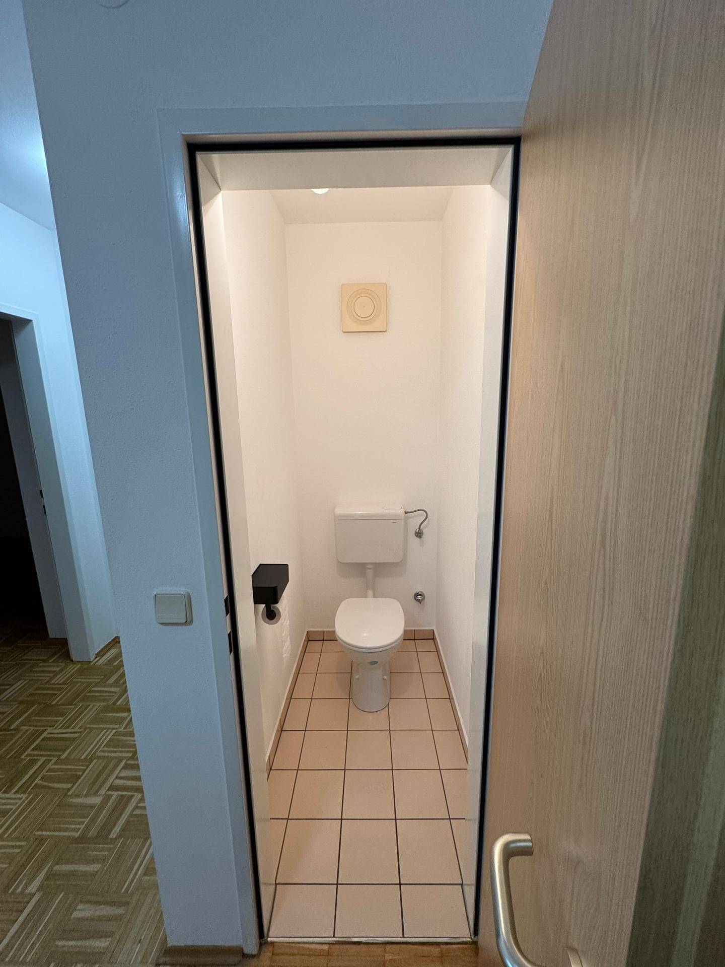 Separates WC mit hellen Bodenfliesen, Toilette und einem Abluftventilator an der Decke.