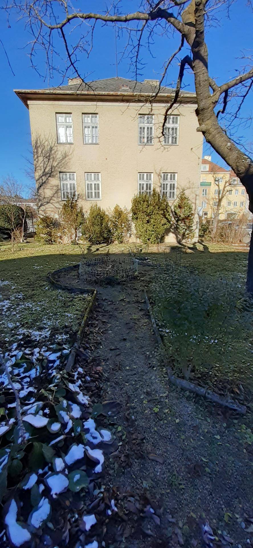 Gartenansicht mit einem Pfad, Bäumen und dem historischen Gebäude im Hintergrund unter blauem Himmel.