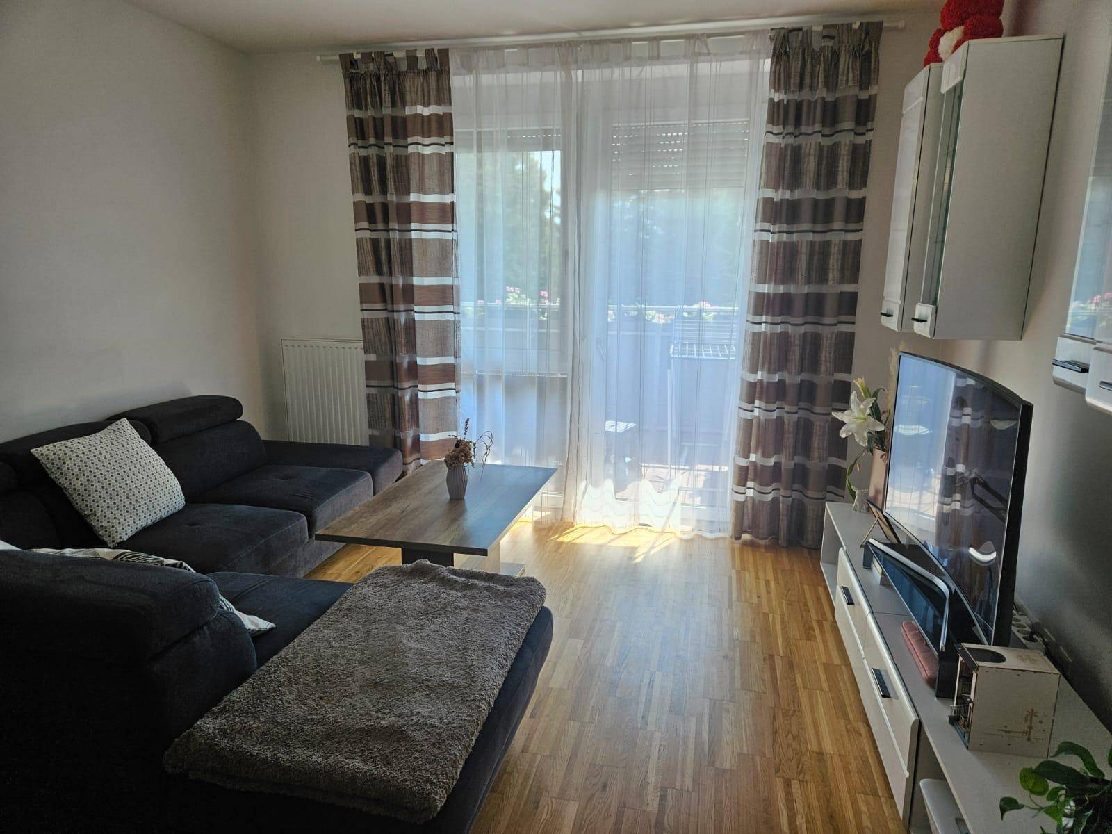 Helles Wohnzimmer mit Sofa, Fernseher und direktem Zugang zum Balkon.