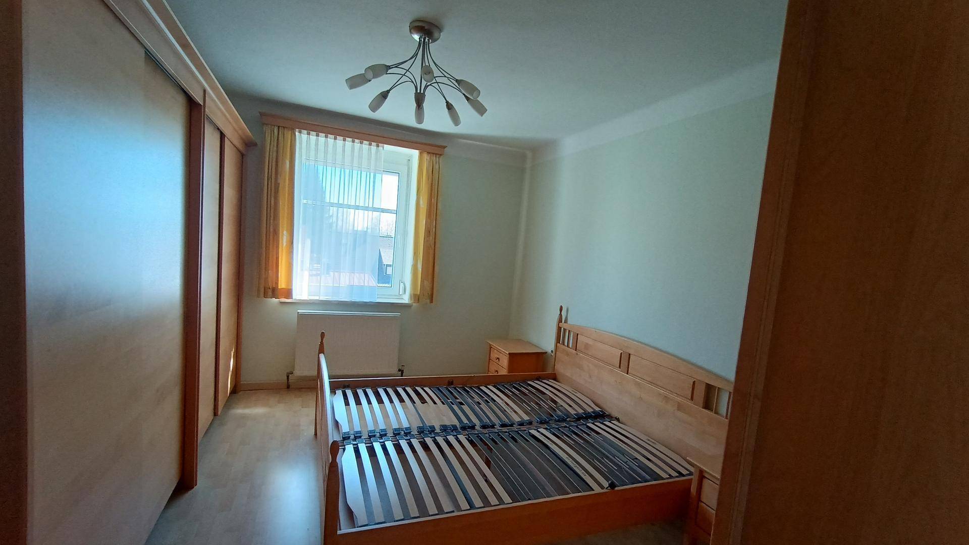 Helles Schlafzimmer mit Holzbett, Kleiderschrank und Fenster mit Vorhängen.