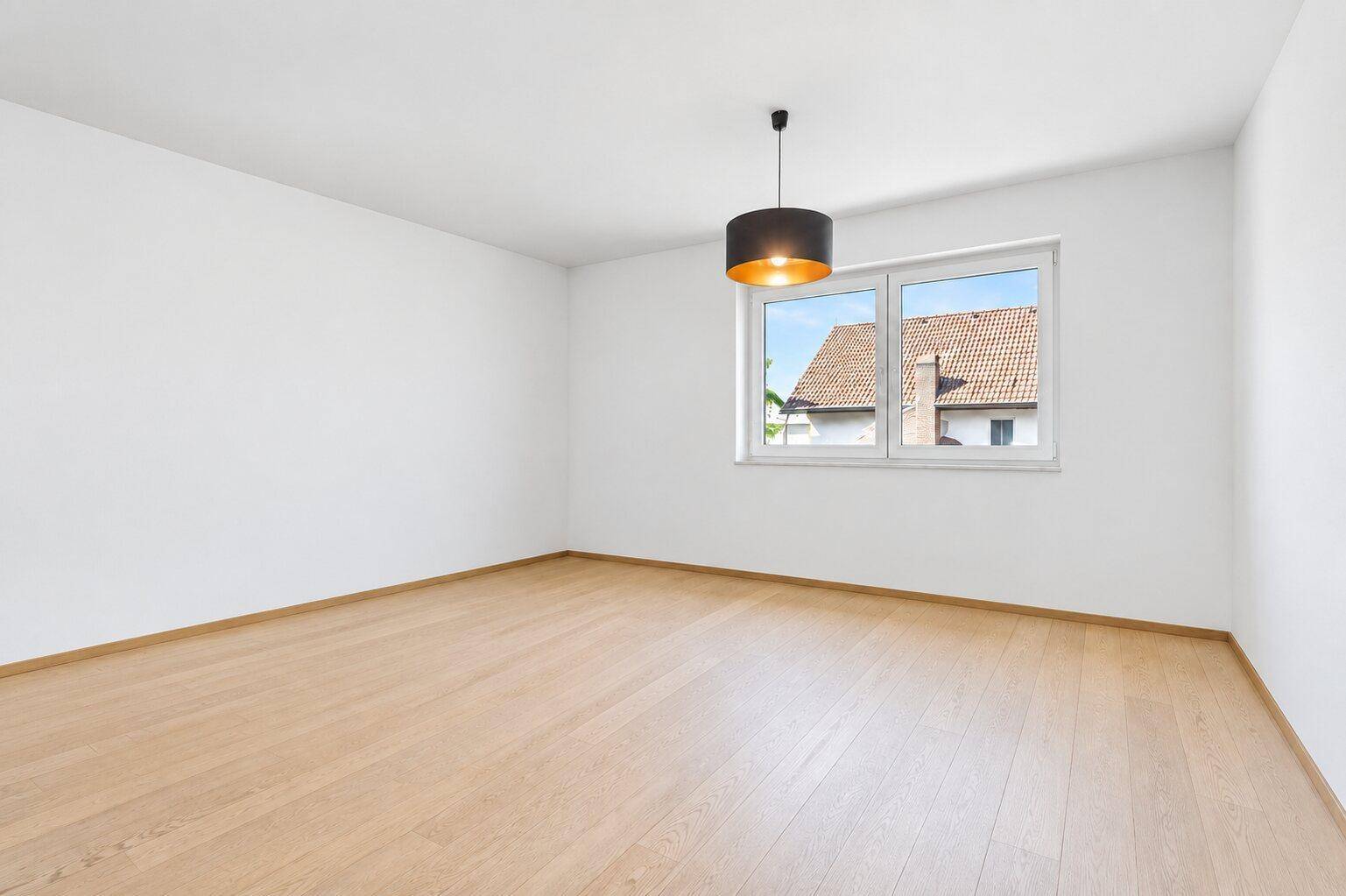 Leeres Zimmer mit hellem Holzfußboden, weißen Wänden und Blick auf die Dächer der Umgebung.