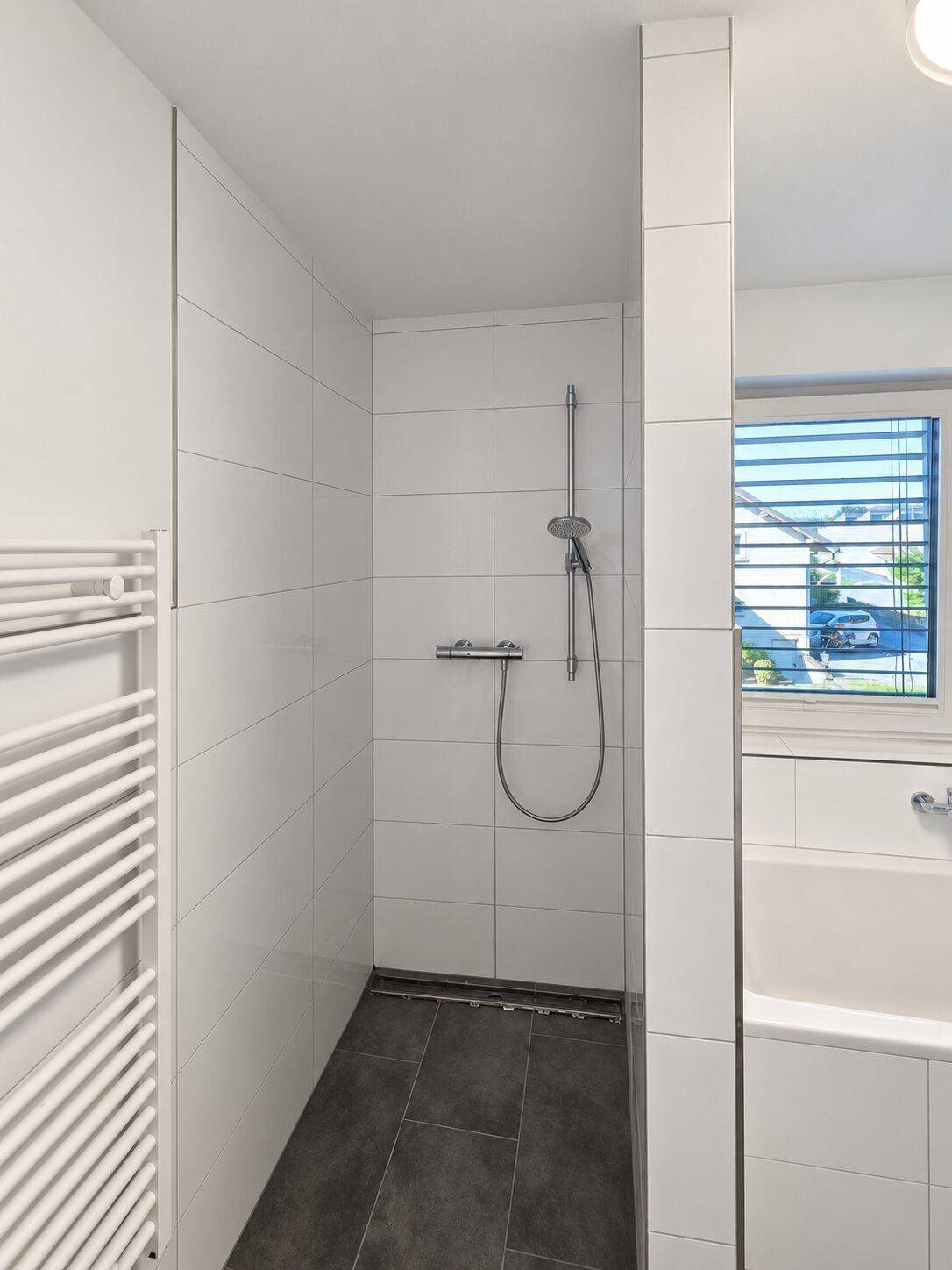 Duschbereich mit weißen Fliesen, dunklem Boden und beheiztem Handtuchhalter im Badezimmer.