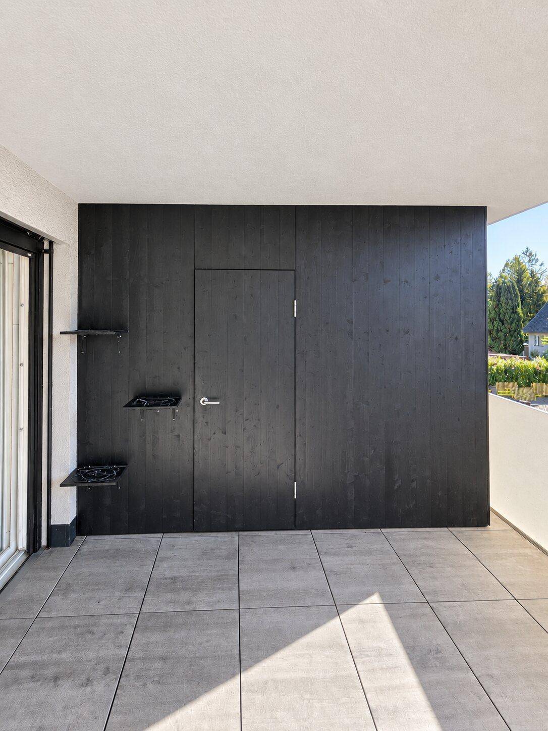 Teil der Terrasse mit einer schwarzen Holzwand, einer Tür und kleinen Ablageflächen.