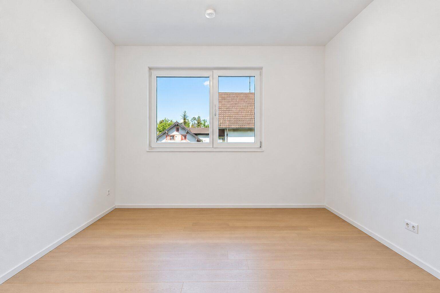 Freundliches, leeres Zimmer mit Parkettboden und Fenster, das einen Blick auf die Umgebung bietet.