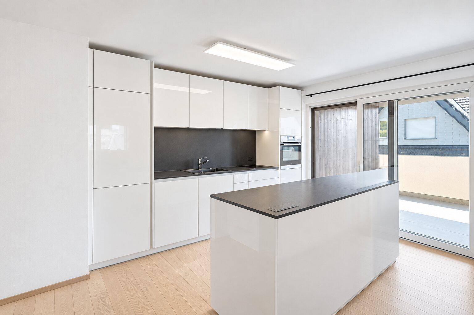 Helle, moderne Küche mit glänzenden weißen Schränken, dunkler Arbeitsplatte und Terrassenzugang.