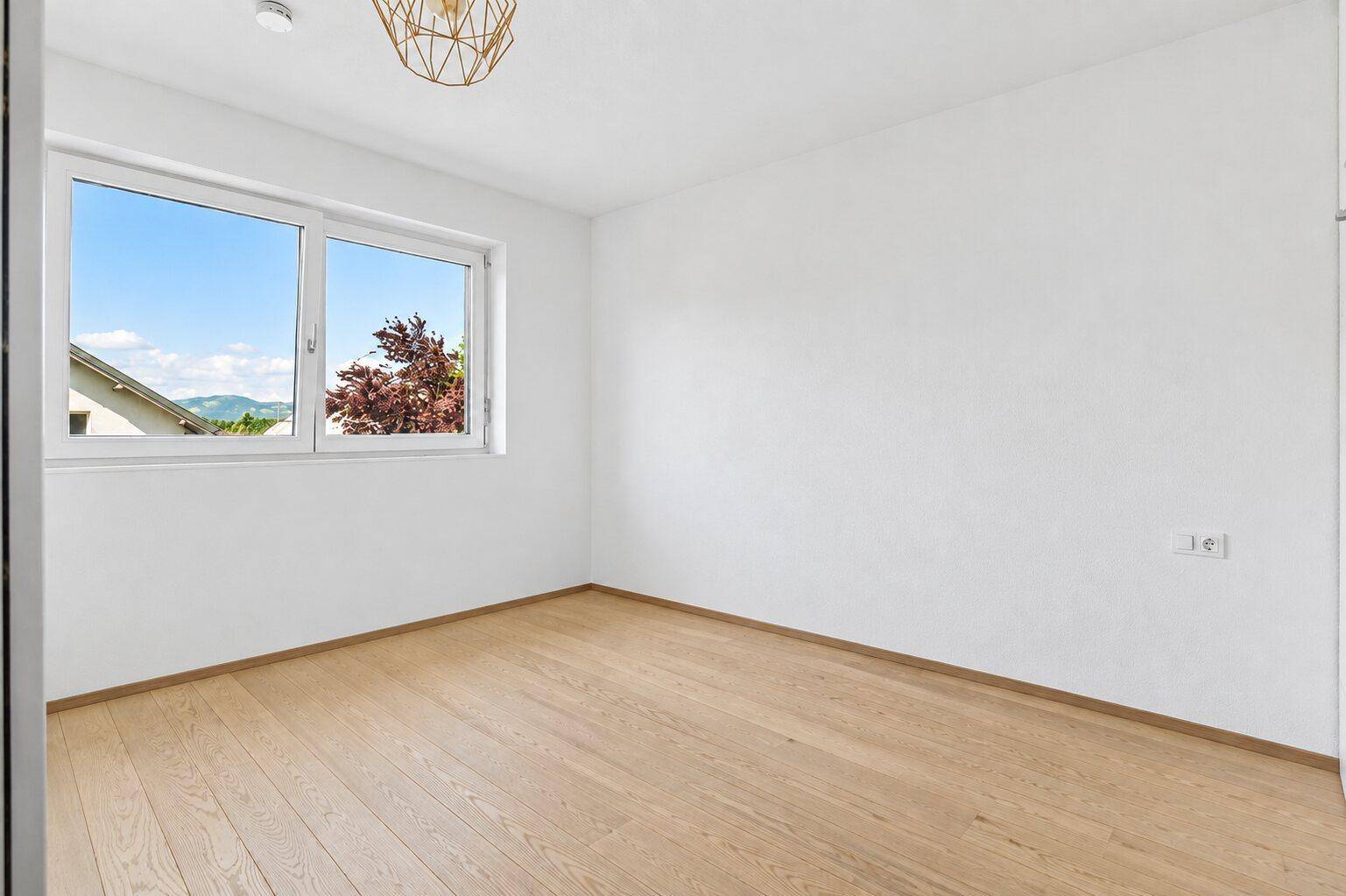 Helles, leeres Zimmer mit Holzfußboden und Fensterblick ins Grüne und auf Nachbarhäuser.