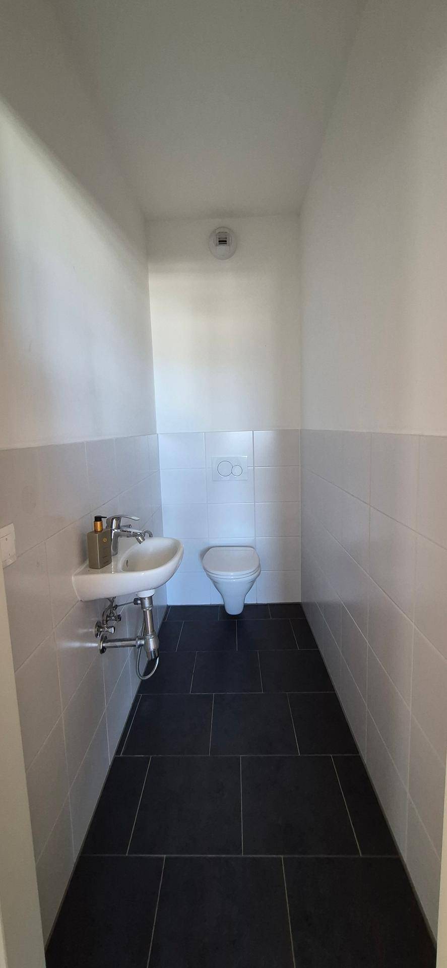 Gäste-WC mit weißer Toilette und Waschbecken, kombiniert mit dunklen Bodenfliesen.