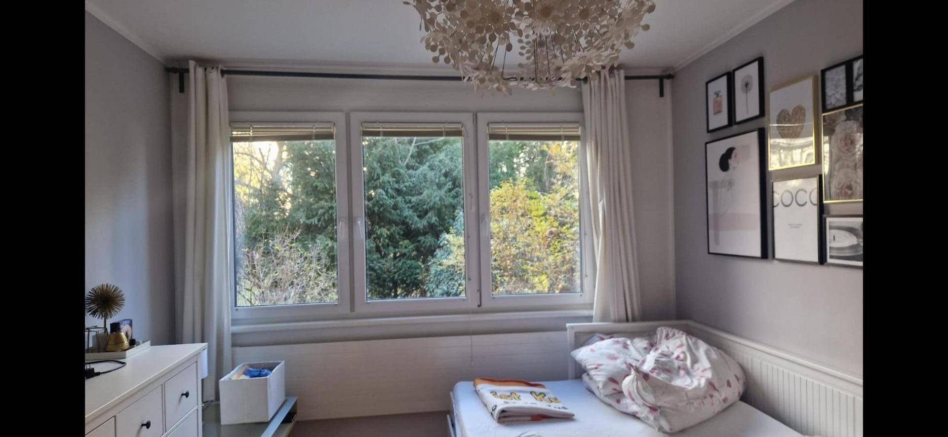 Helles Schlafzimmer mit Einzelbett, Kommode und Blick ins Grüne durch große Fenster.