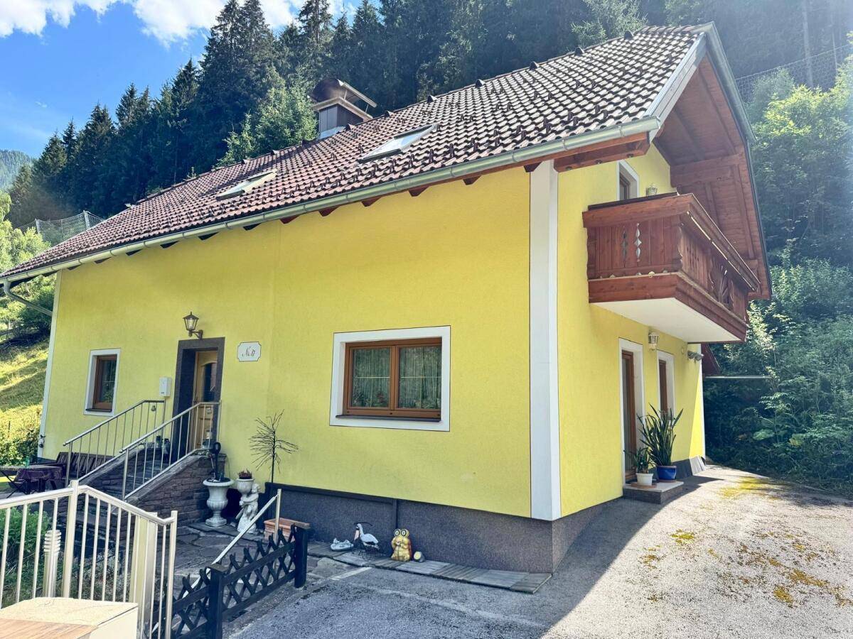 Nahaufnahme der gelben Hausfassade mit Holzbalkon, Eingangsbereich und Zugang zur Dachterrasse.