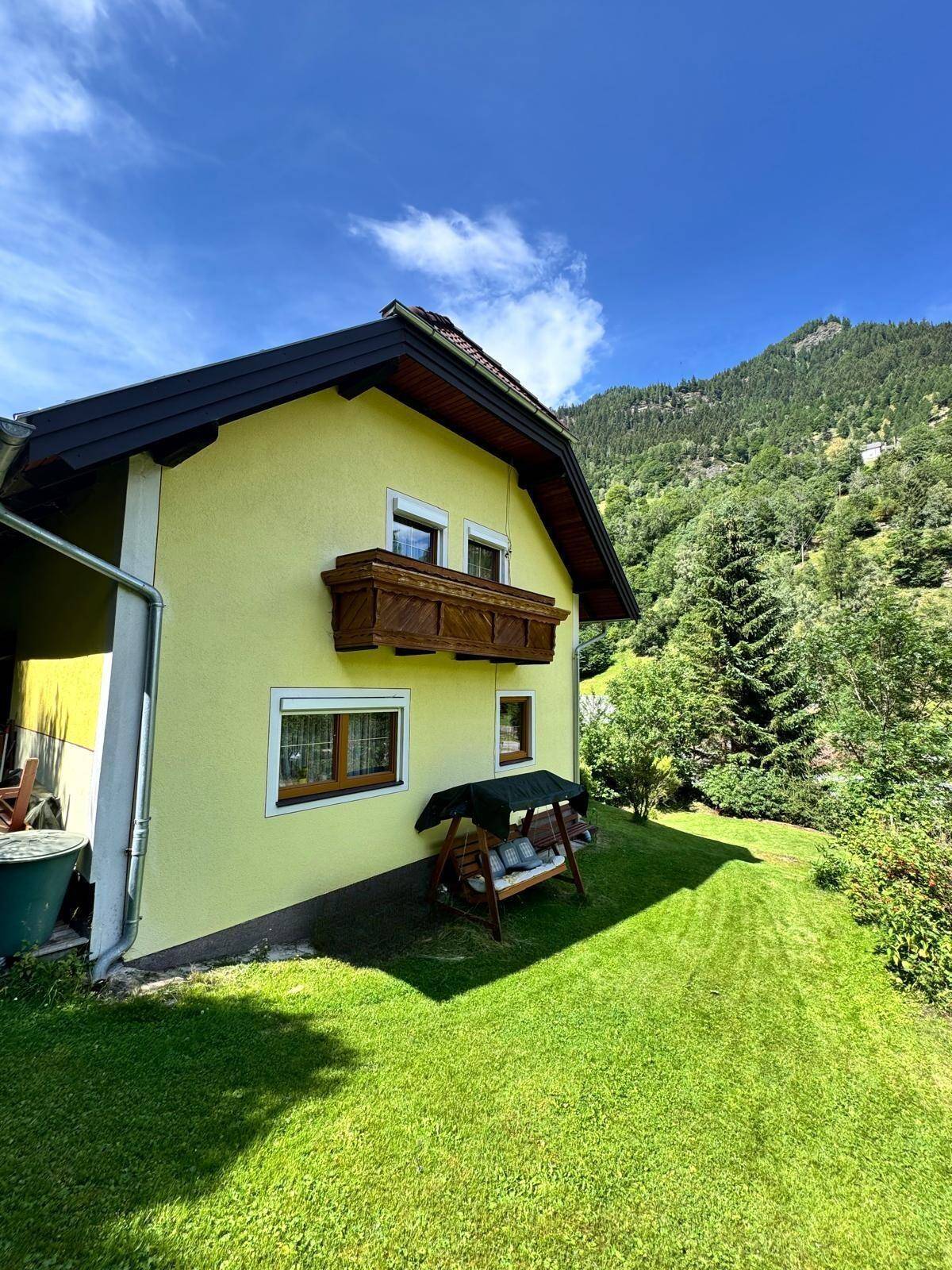 Seitenansicht des Hauses mit Holzbalkon, Gartenschaukel und weitläufiger grüner Rasenfläche.
